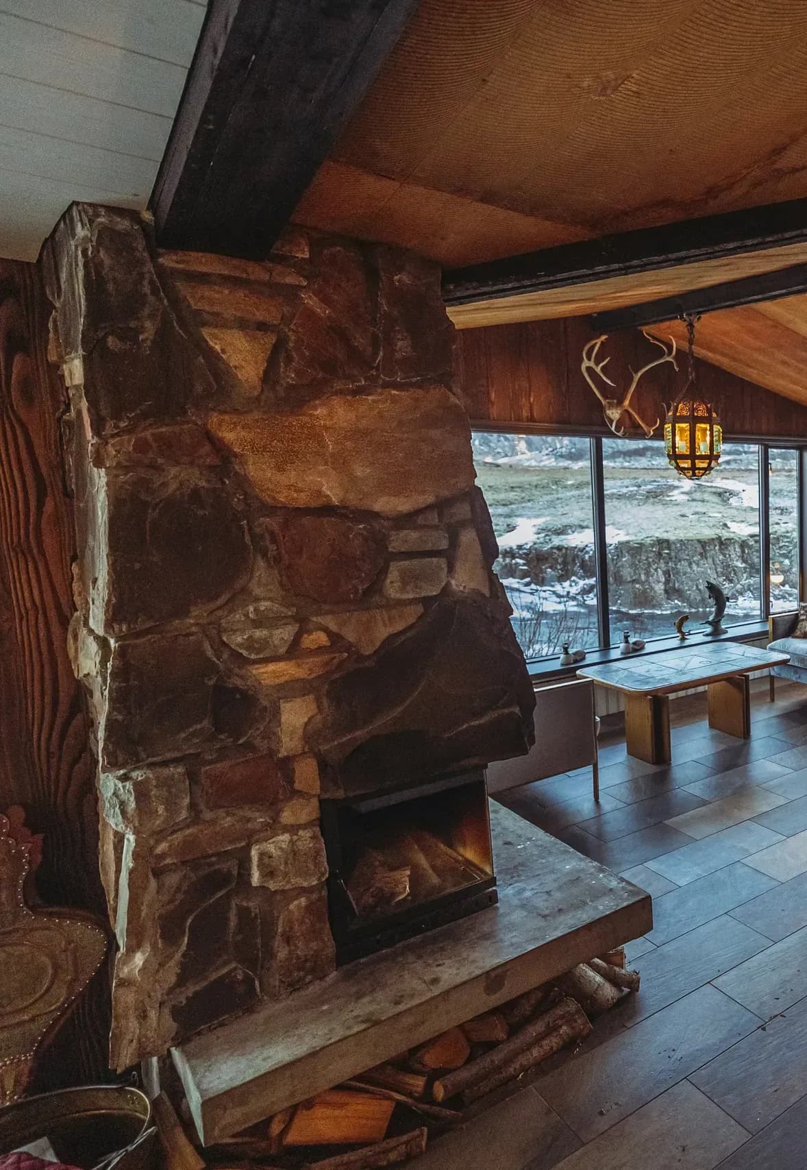 A stone fireplace burning in the timber lounge under an antler lantern