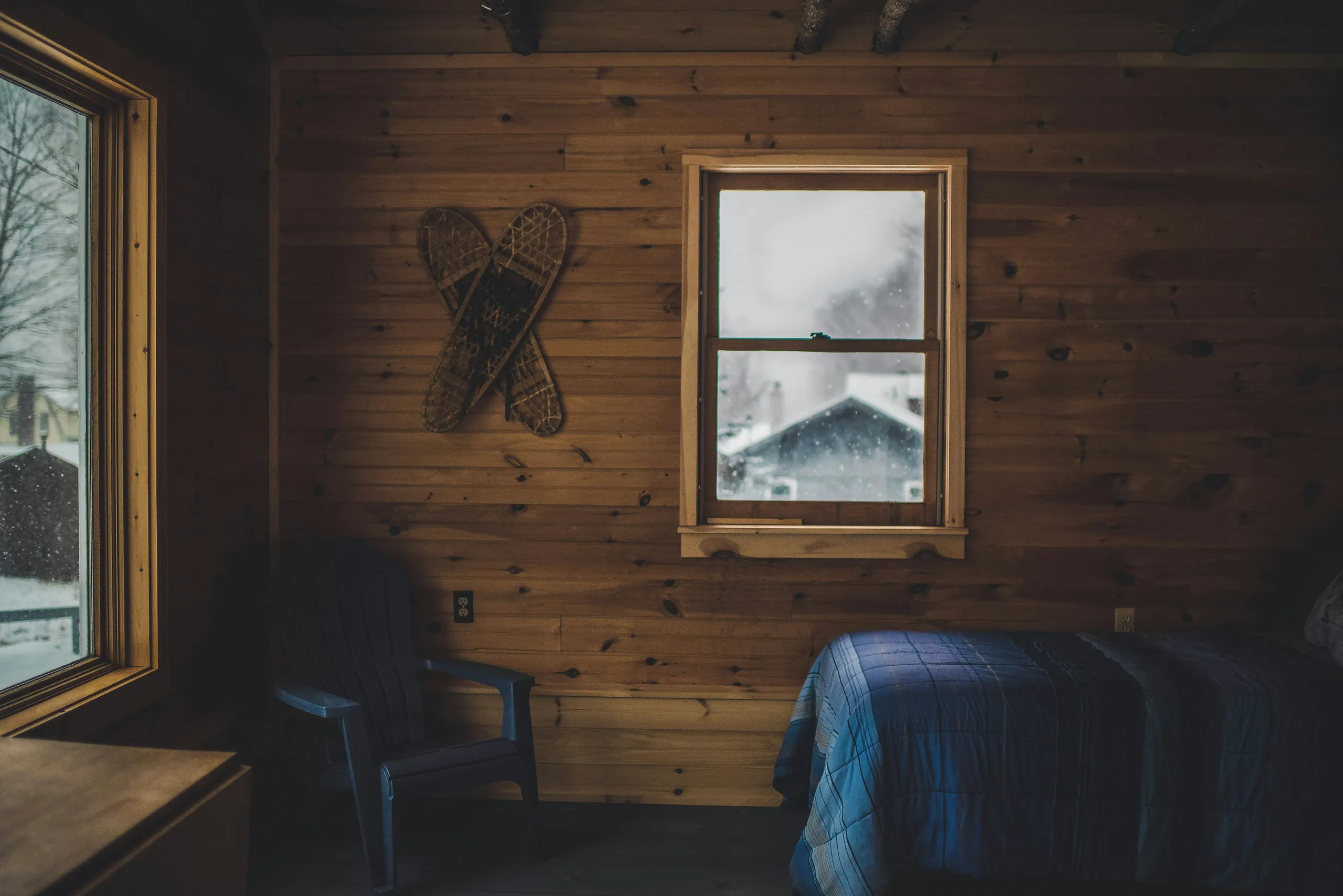Pine-boarded cabin room with old snowshoes hung on the wall and snow falling past a sash window