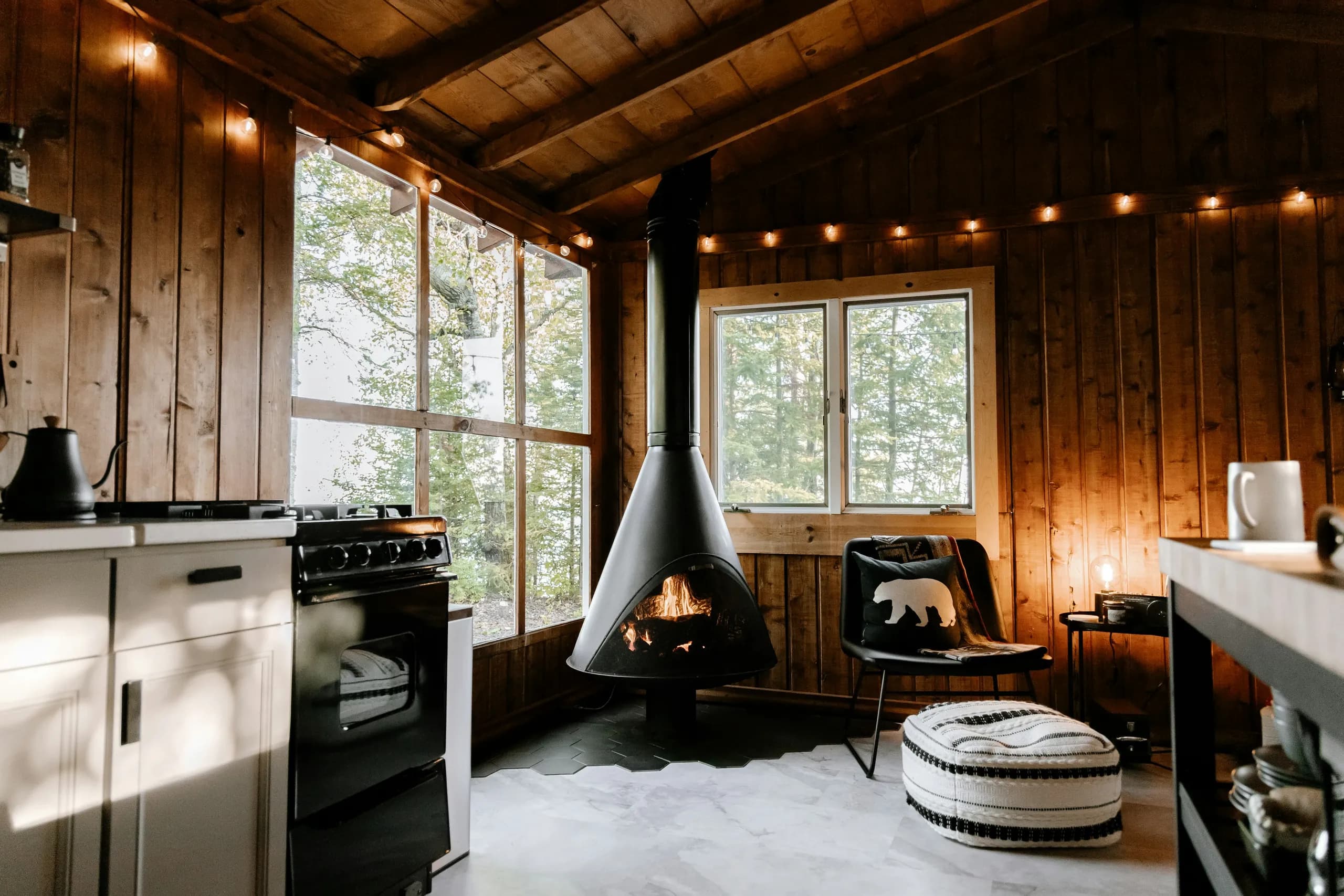 Timber-walled cabin sitting room with a black conical wood stove alight, a small kitchen corner and windows onto the pines