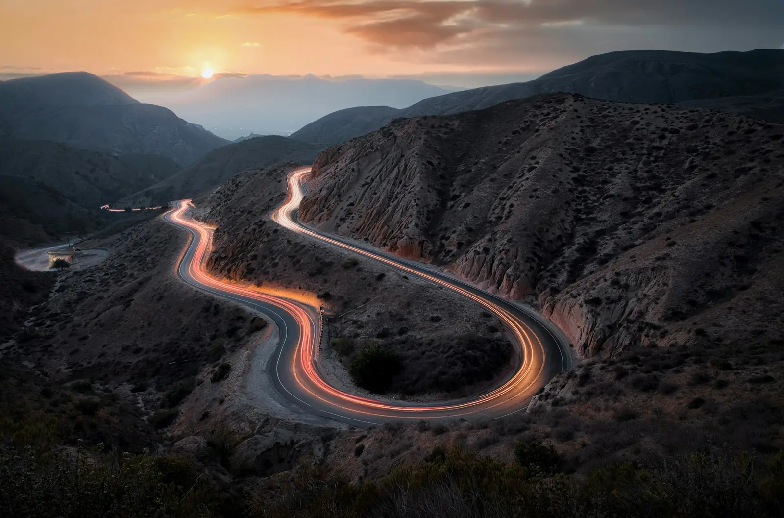 A mountain switchback traced by ember light trails at dusk, ridgelines fading behind