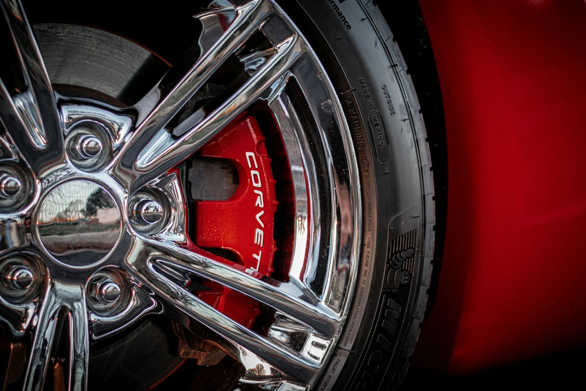 A red Corvette brake caliper behind a polished chrome wheel, the tyre sidewall lettering sharp