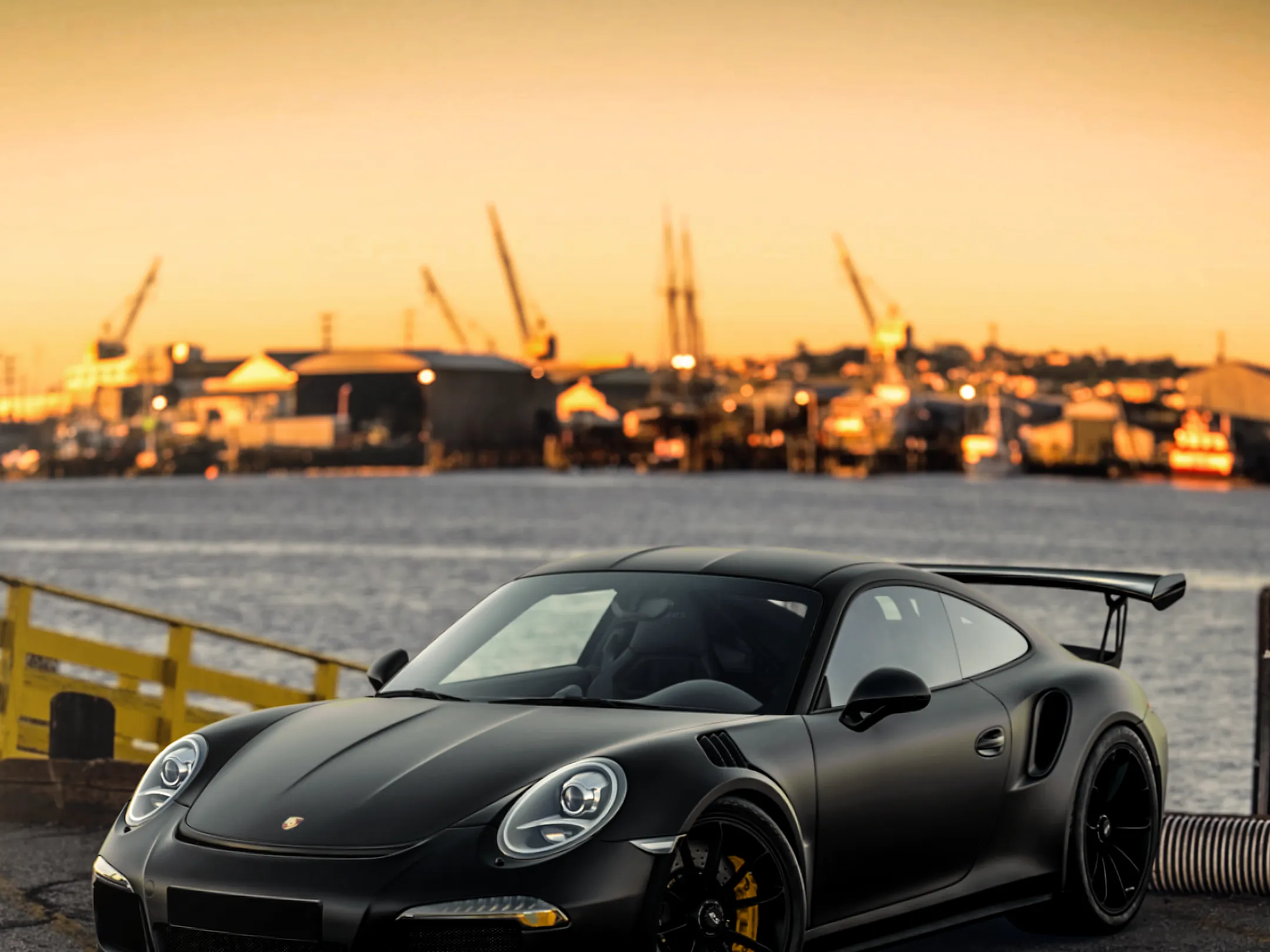 A matte black Porsche 911 parked on a quayside at sunrise, the sky burning orange behind the cranes