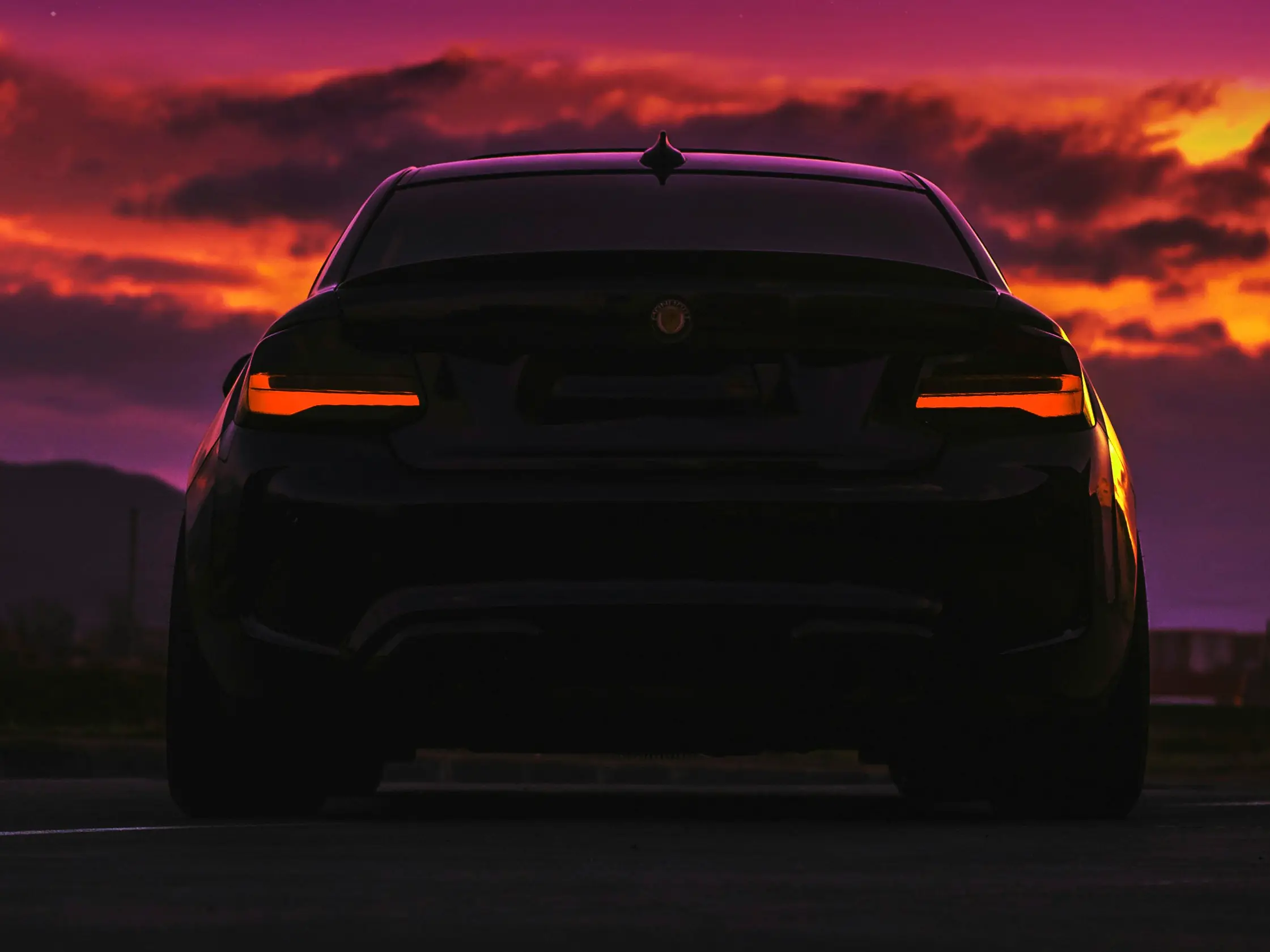 The rear of a dark coupe silhouetted against a burning orange and violet sunset, its tail lamps lit