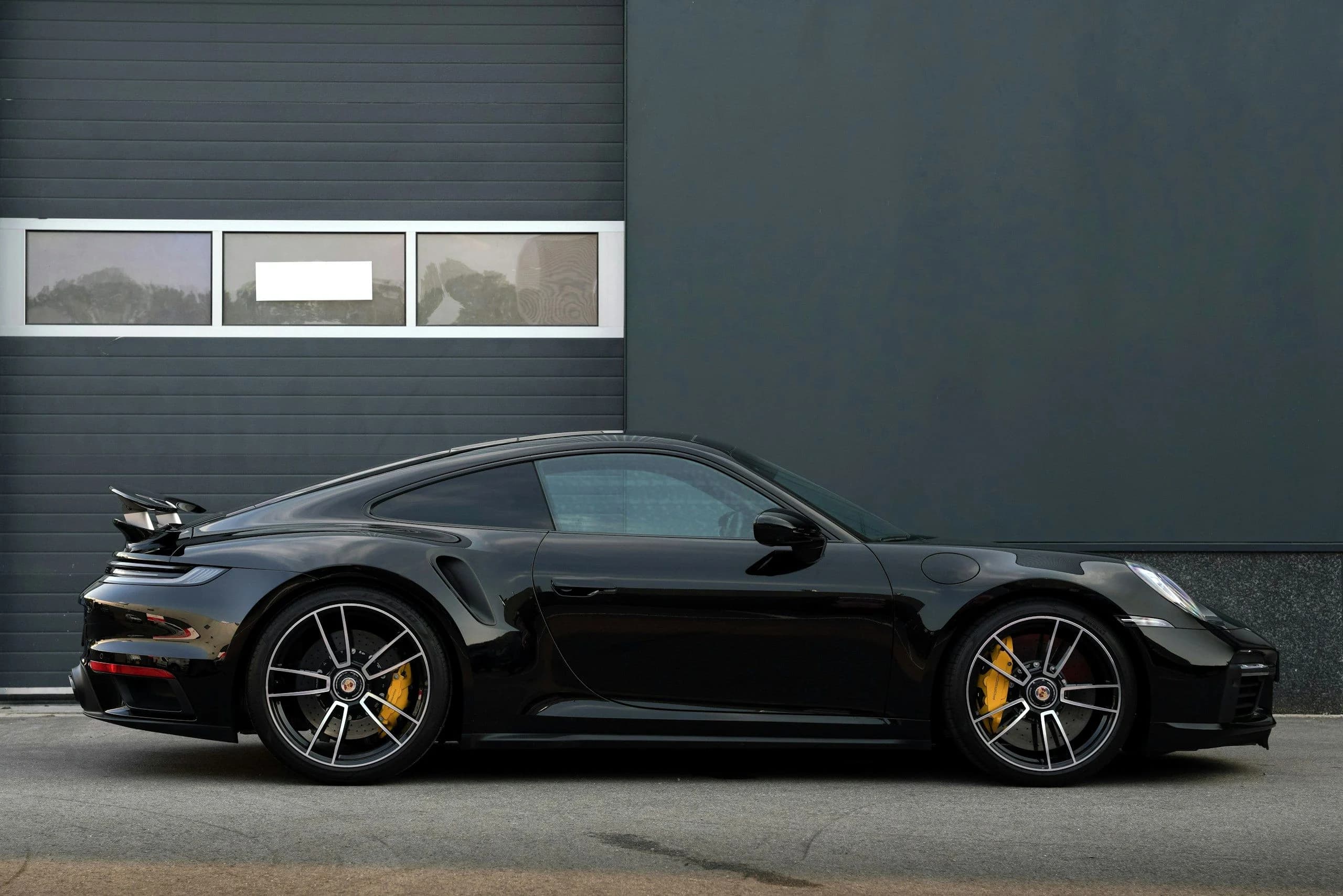 A black Porsche 911 Turbo S photographed in full profile against a plain grey wall, yellow ceramic brake calipers behind the wheels