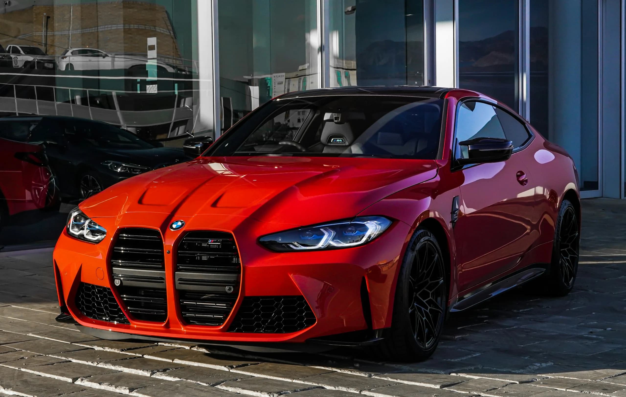 A red BMW M4 Competition in front-three-quarter view outside a lit showroom, its tall kidney grille and M badge in full view