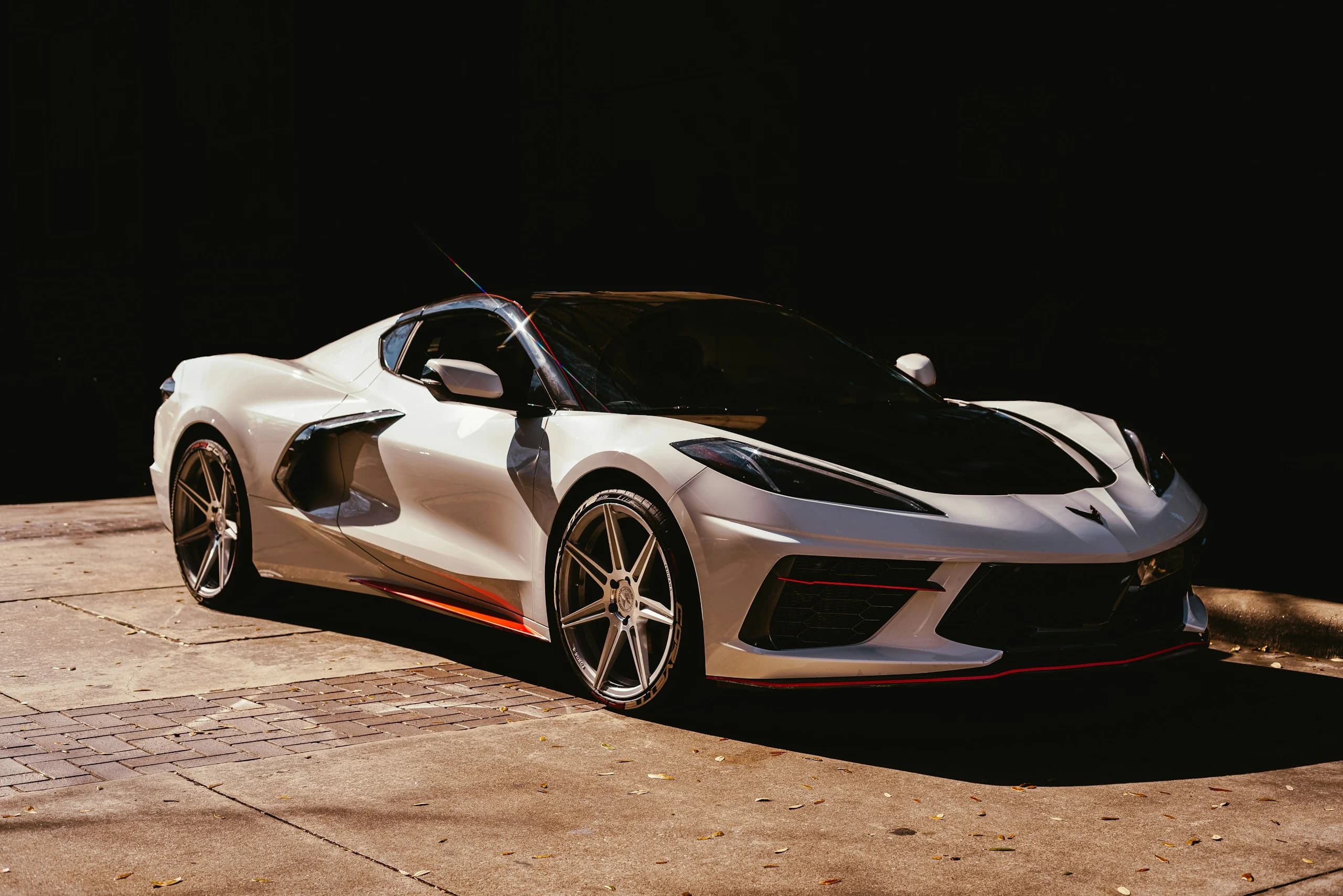 A white mid-engined Chevrolet Corvette Stingray lit hard against a black background, the crossed-flags badge on its nose