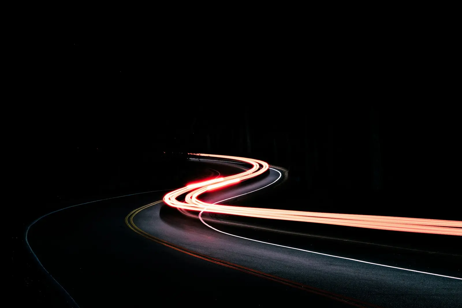 Ember tail-light streaks drawing an S-curve down an unlit road, long exposure