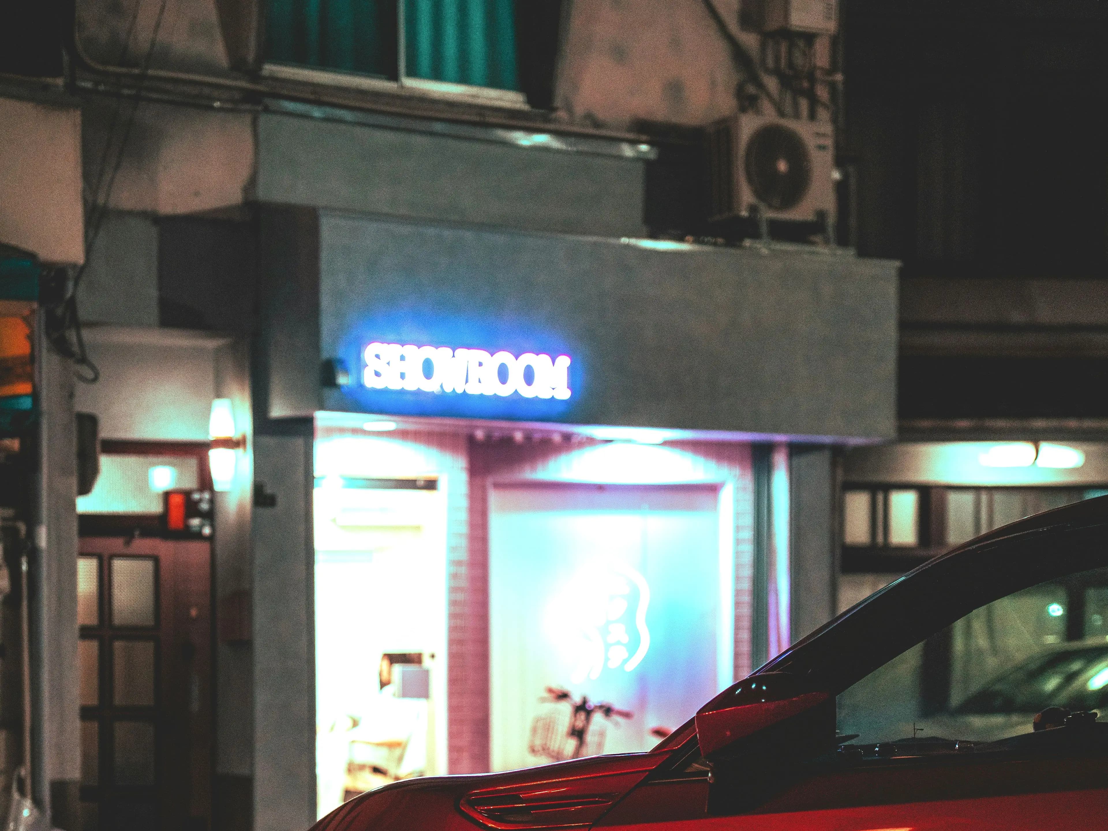 A showroom sign burning above a wet street at night, the nose of a red car at the kerb