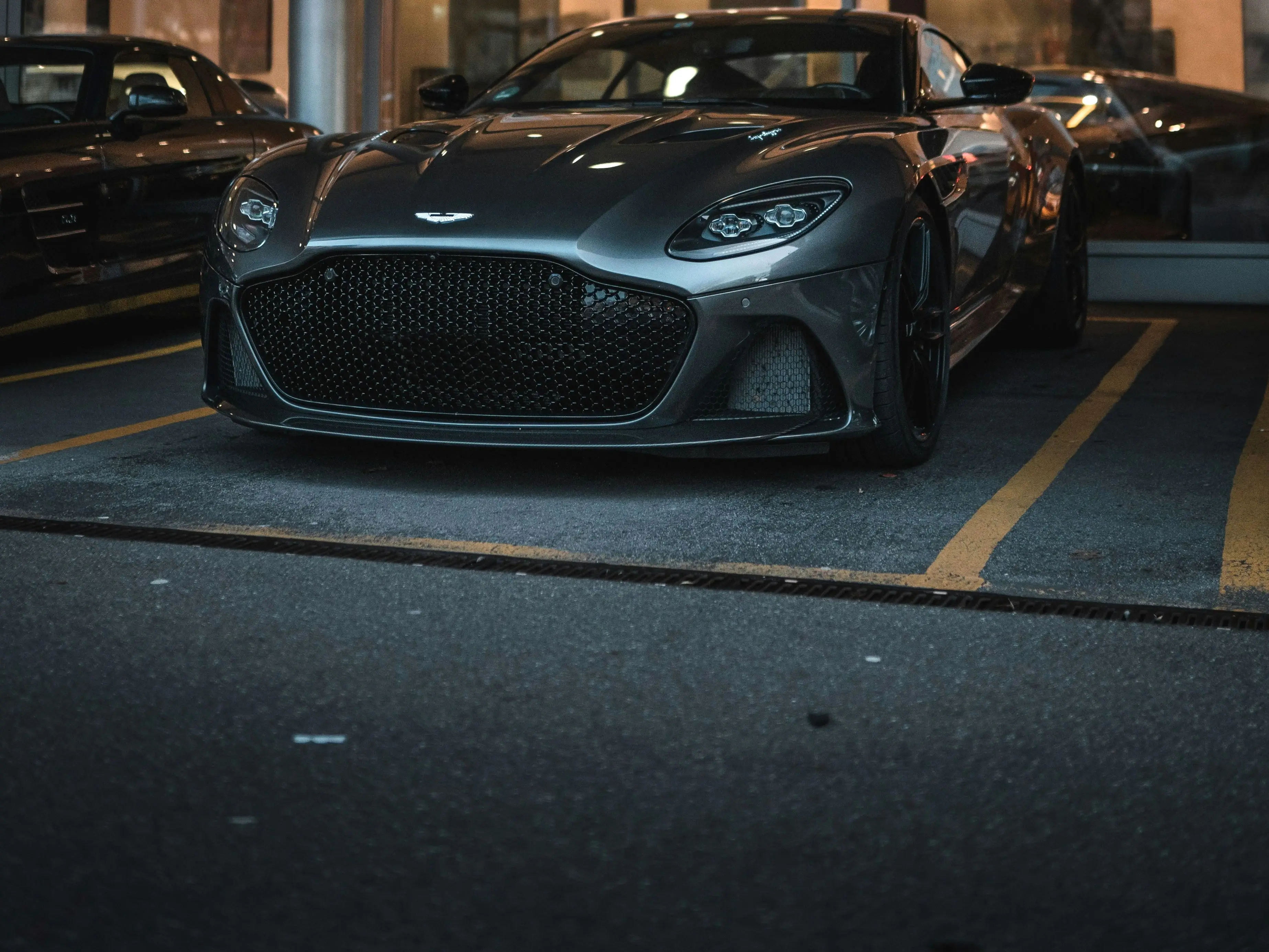 A silver Aston Martin standing on the forecourt outside a lit showroom at dusk