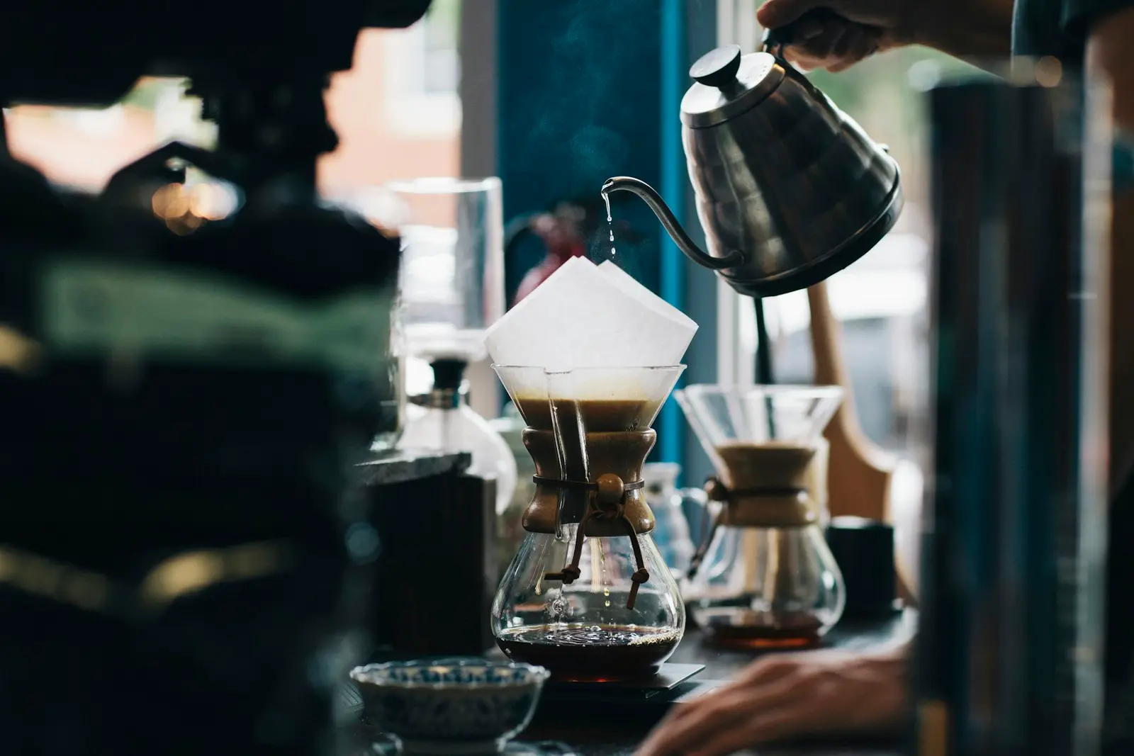 Barista pouring hot water from a gooseneck kettle into a Chemex brewer by a bright window