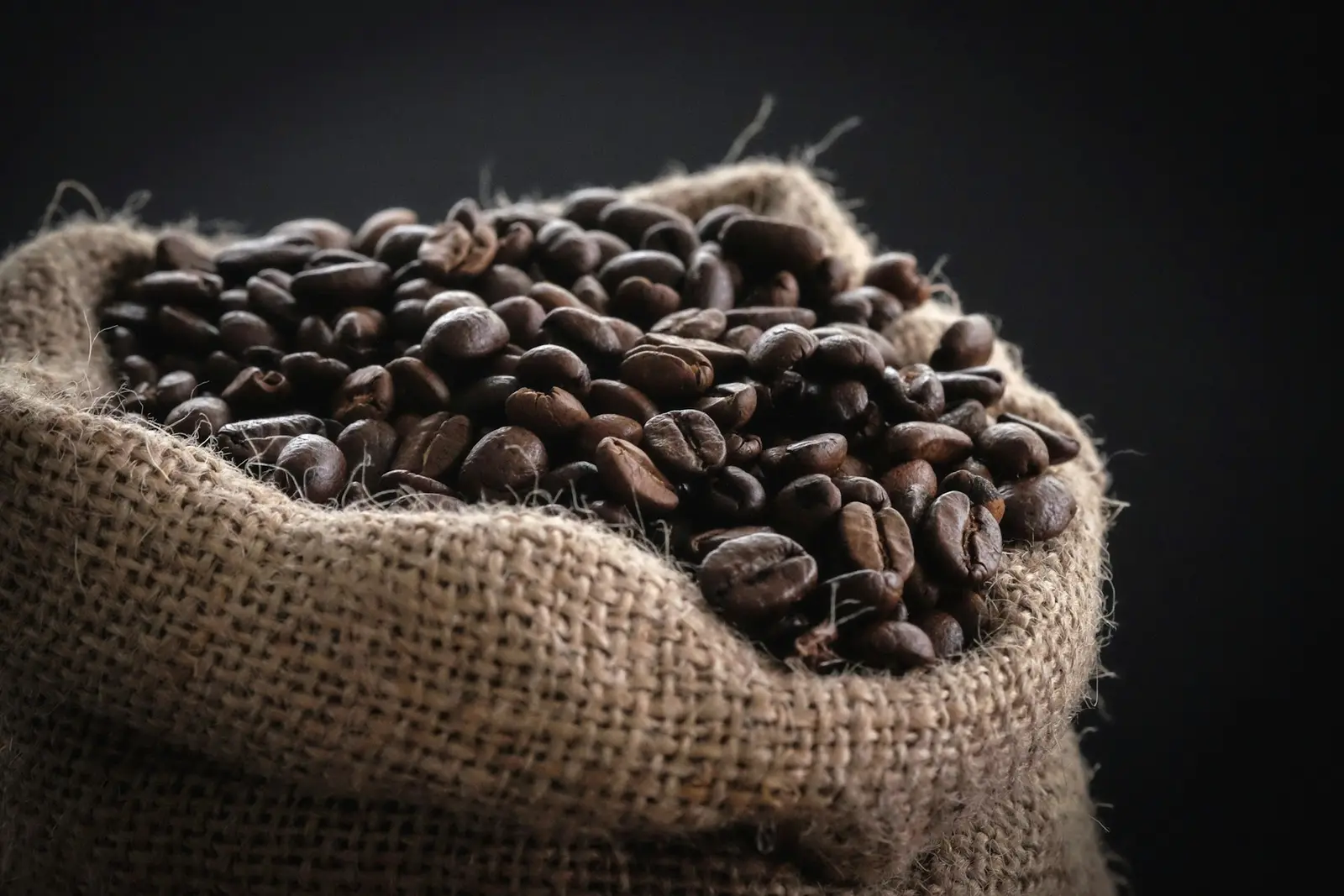 Burlap sack overflowing with dark roasted coffee beans against a black backdrop