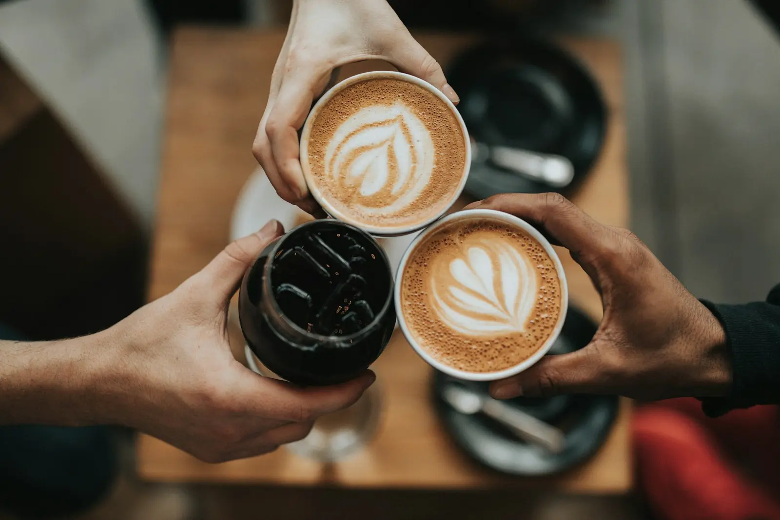 Three friends raising a latte, a cappuccino and a cold brew over a wooden table