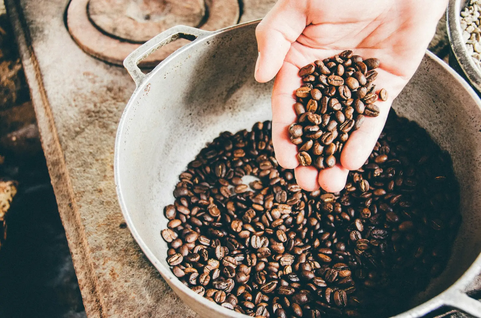 Hand lifting a scoop of roasted coffee beans from a metal pan on an old stove