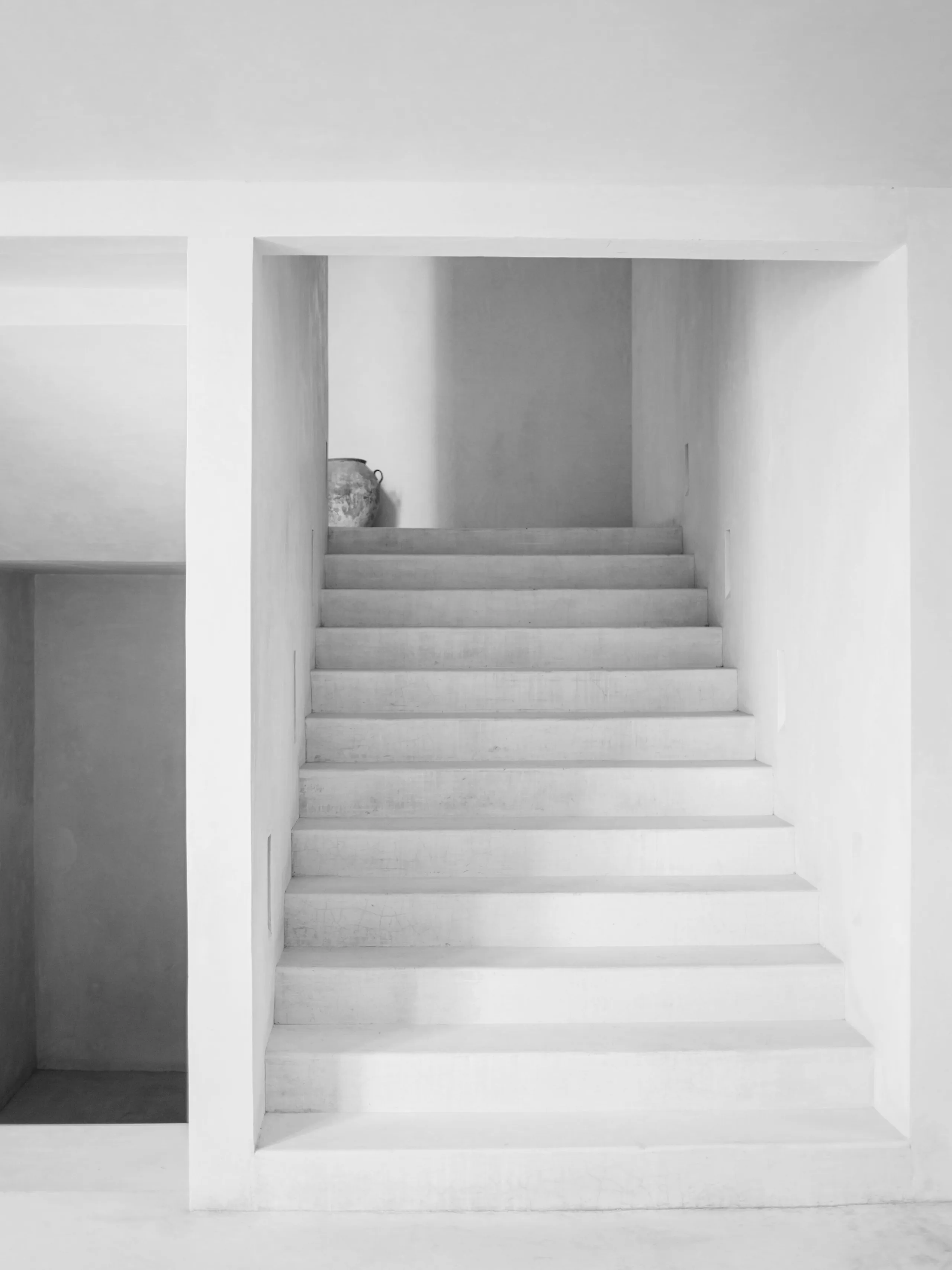 Monochrome photograph of a white plastered stair rising through a square reveal, a single vessel standing on the top landing