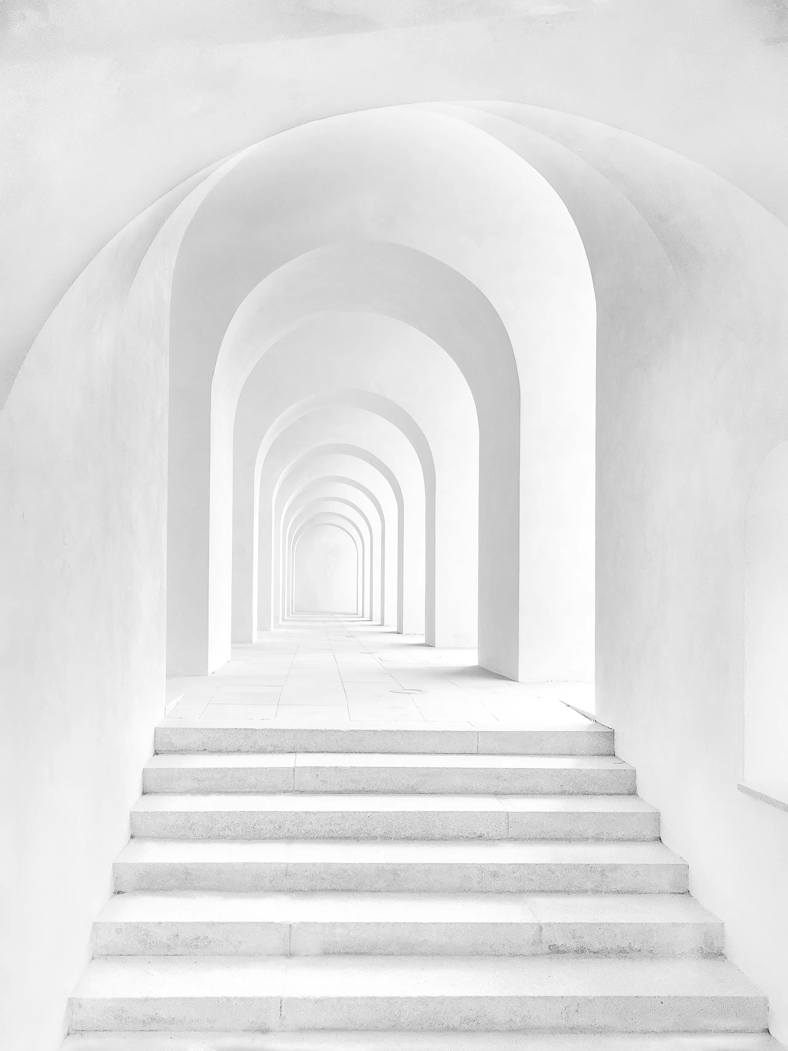 Monochrome photograph of a white vaulted arcade: a long run of repeating arches receding into the distance above a flight of shallow steps