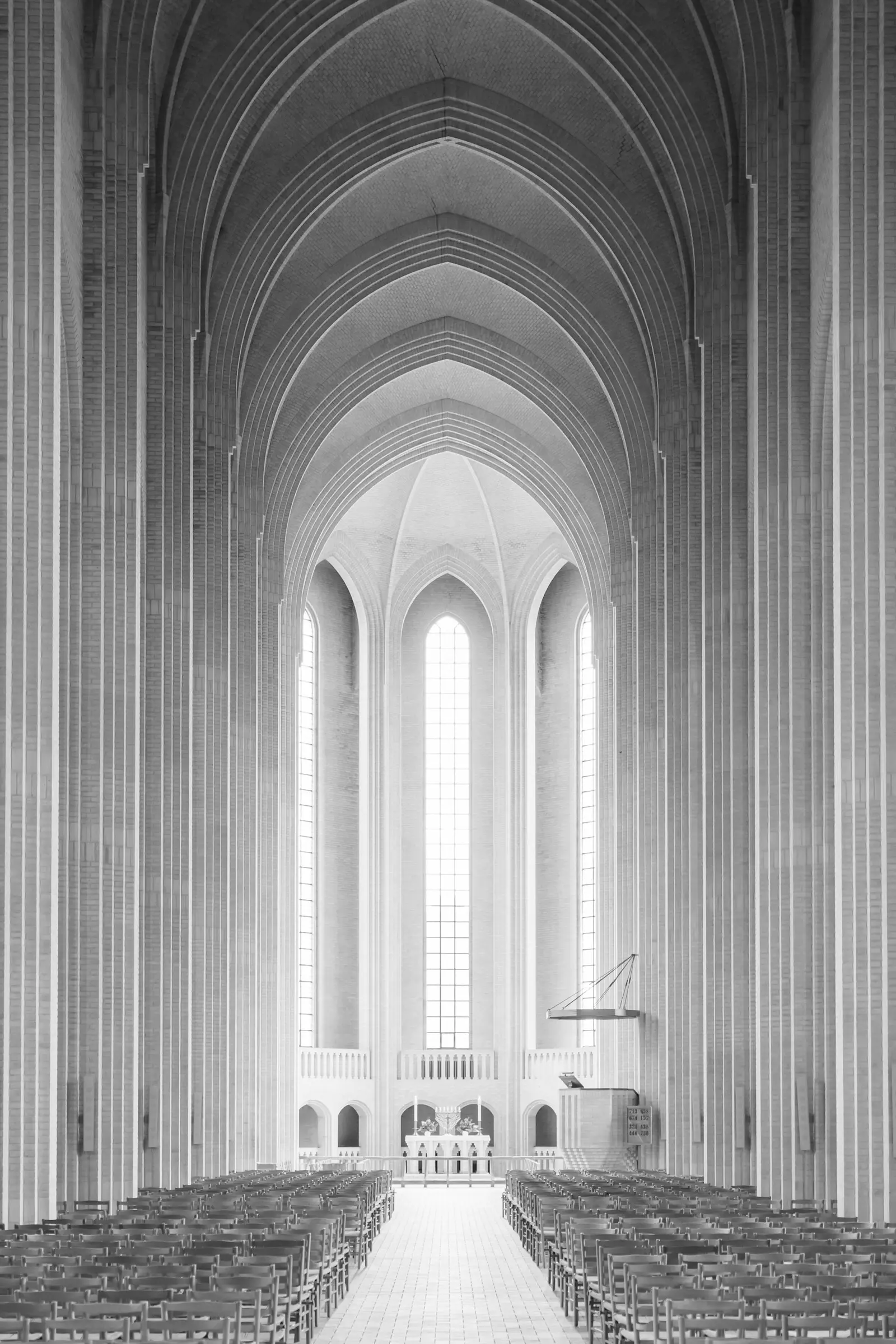 Monochrome photograph of a vast brick church nave with ribbed vaulting, tall arched windows and rows of chairs facing the altar