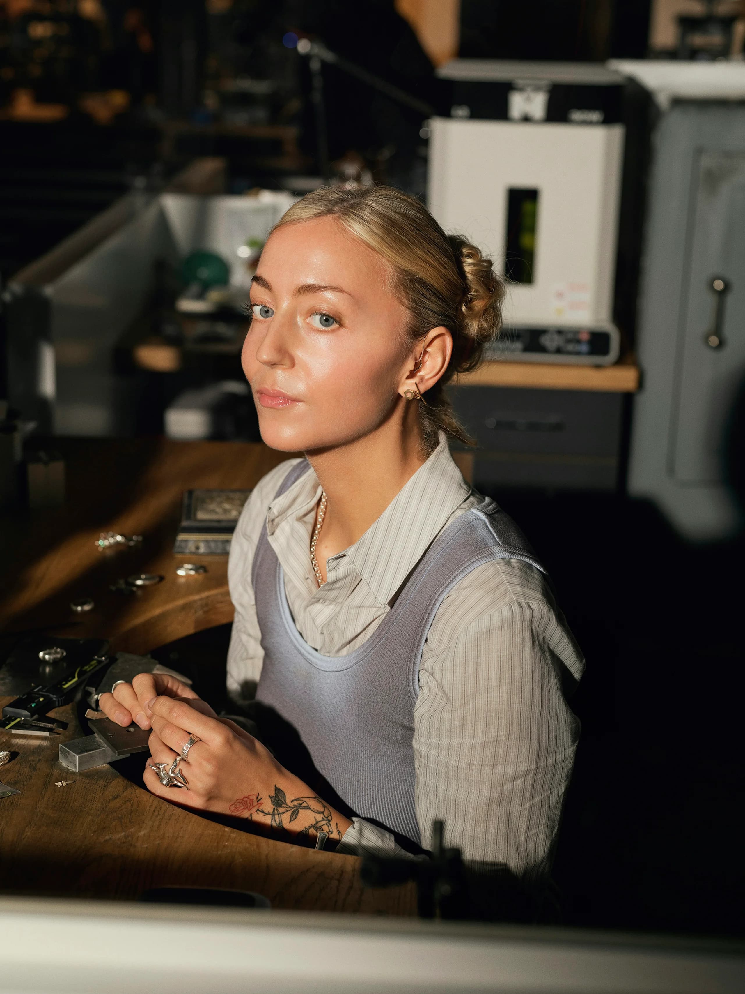 A jeweller sitting at the setting bench in warm evening light, rings on her own fingers, tools laid out in front of her