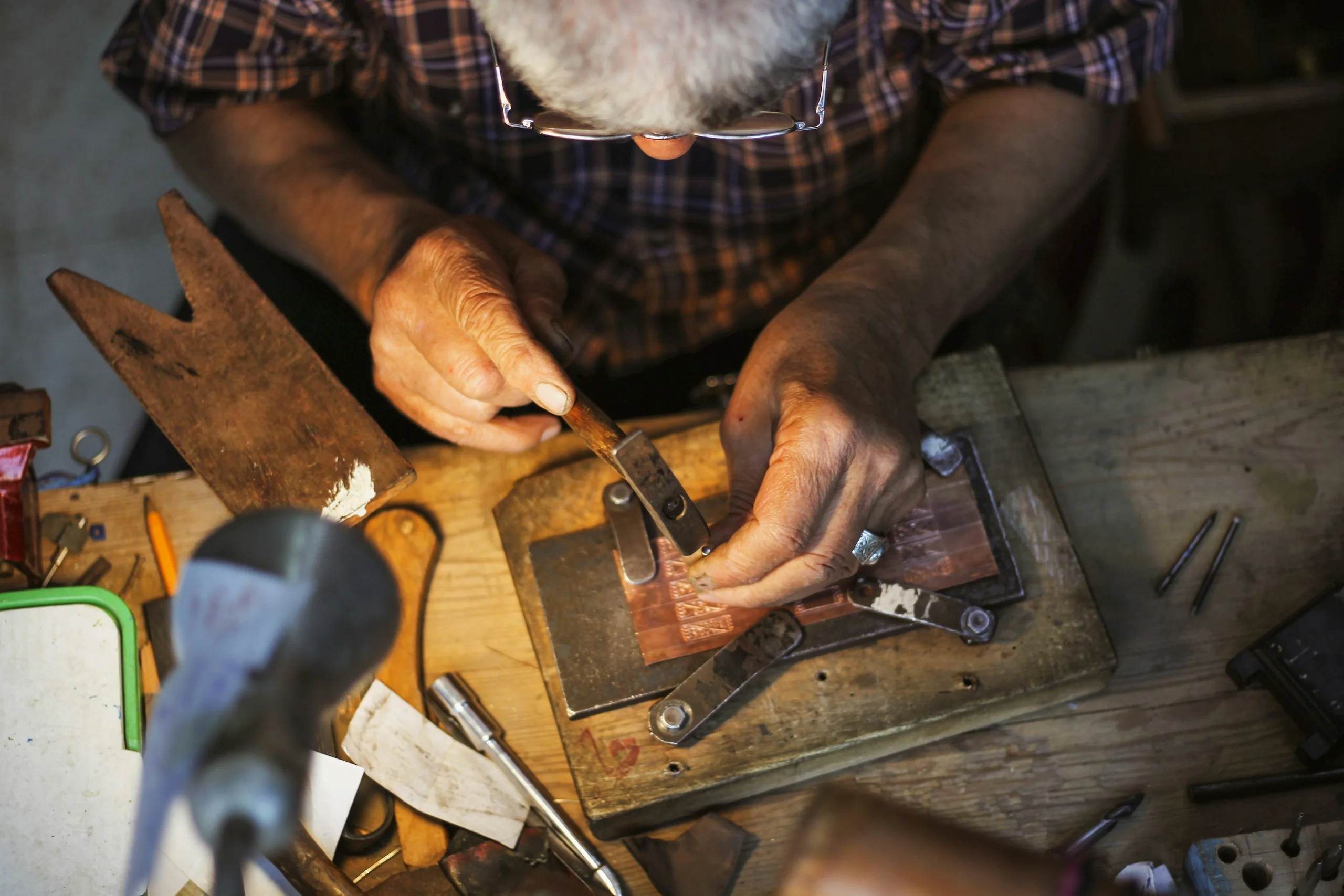A goldsmith cutting a design into a flat copper plate clamped to a bench block, gravers and hammers around him