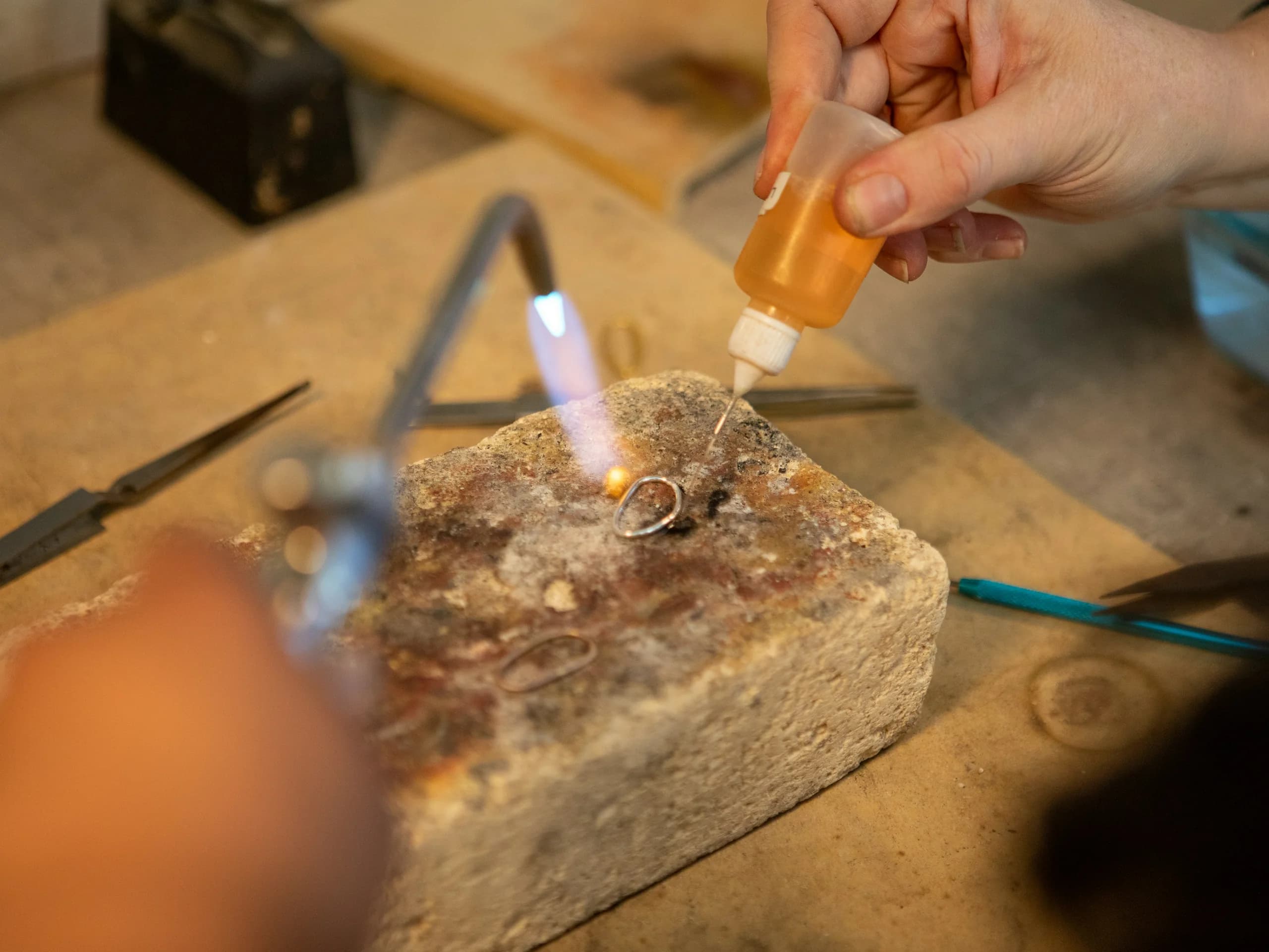 A hand laying flux from a dropper bottle onto a ring on the soldering block while the torch burns