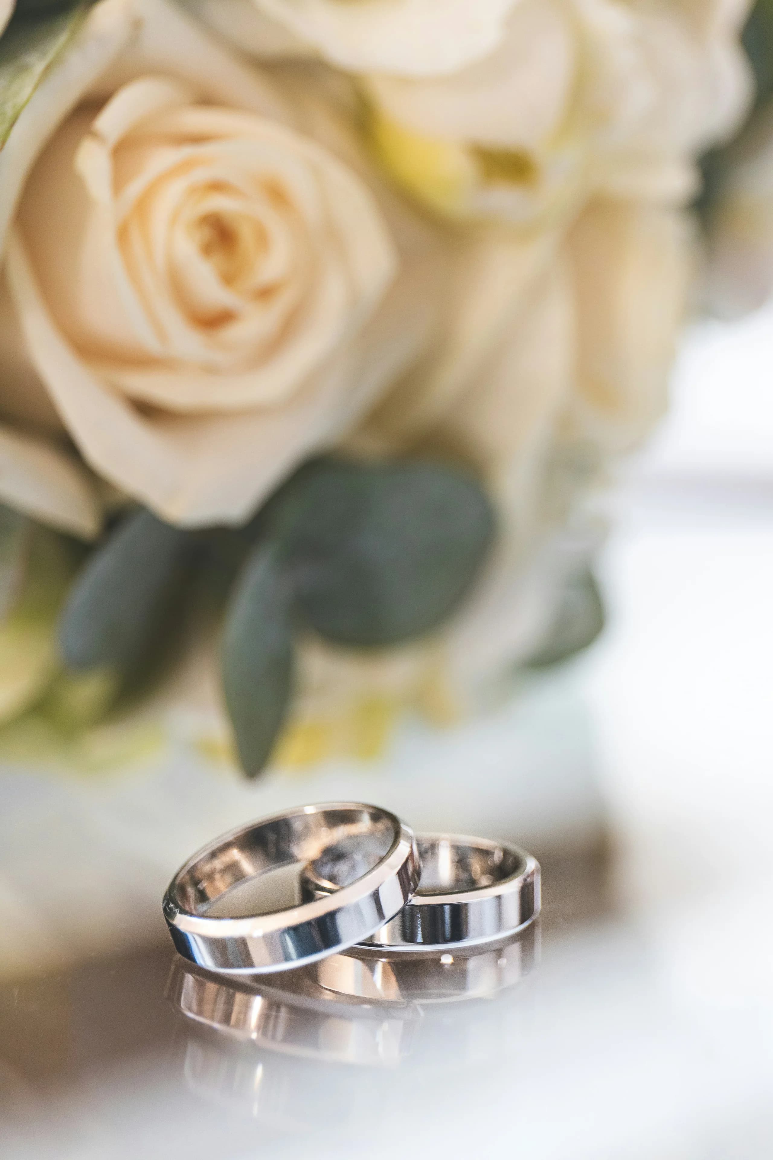 Two plain flat-section bands in white metal resting on a polished surface in front of a cream rose bouquet