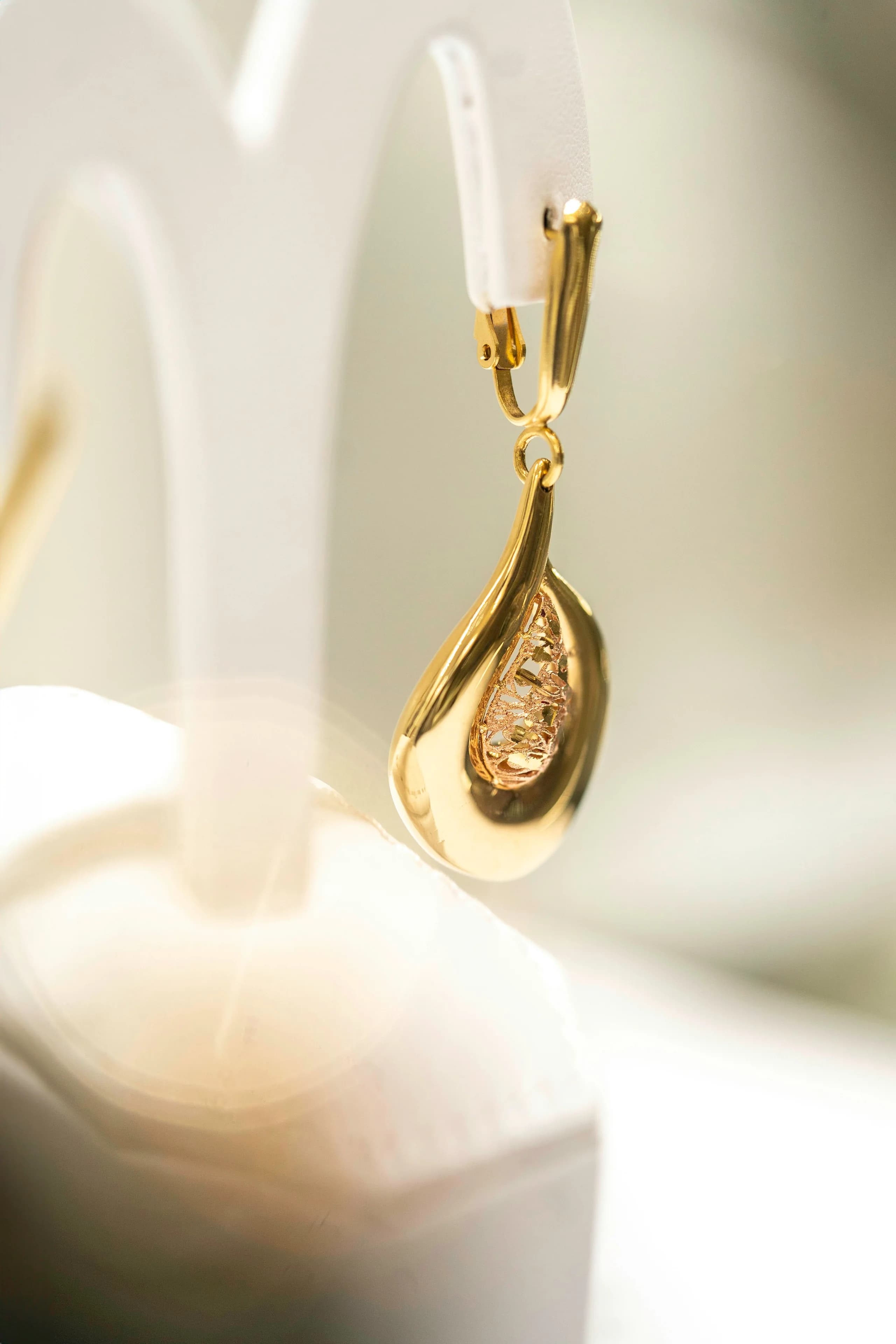 A smooth polished gold teardrop on a lever-back hook, hanging from a white display stand in soft daylight