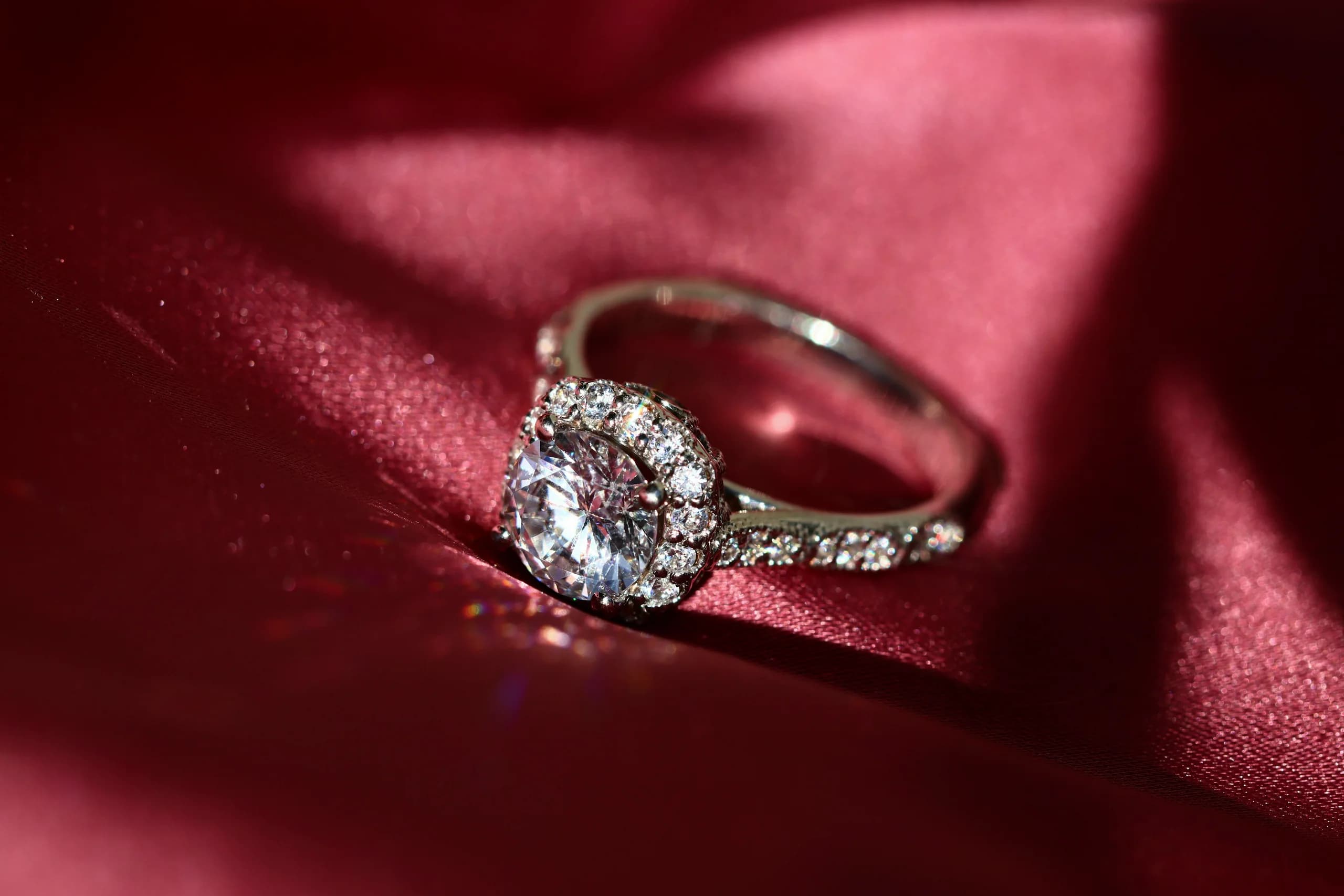 A round brilliant in a claw-set halo, white metal, pavé down both shoulders, lying on deep red satin