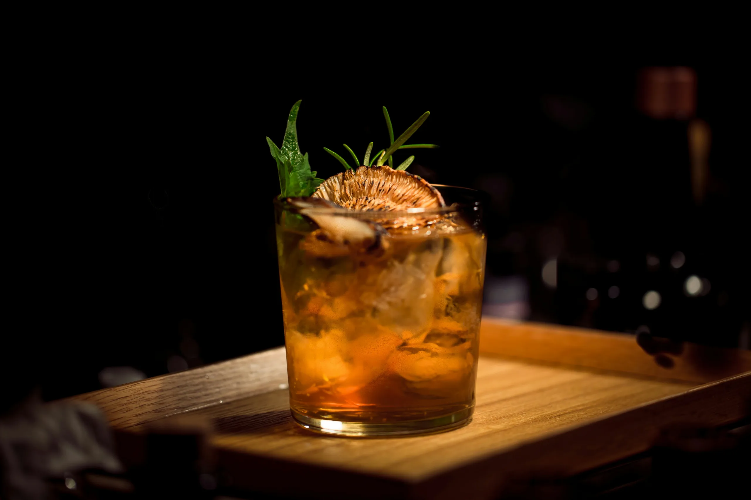 An amber non-alcoholic drink over ice with rosemary and dried citrus on a wooden board
