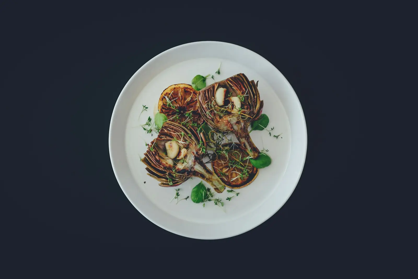 Roasted artichoke halves with herbs and charred lemon on a white plate against a dark table