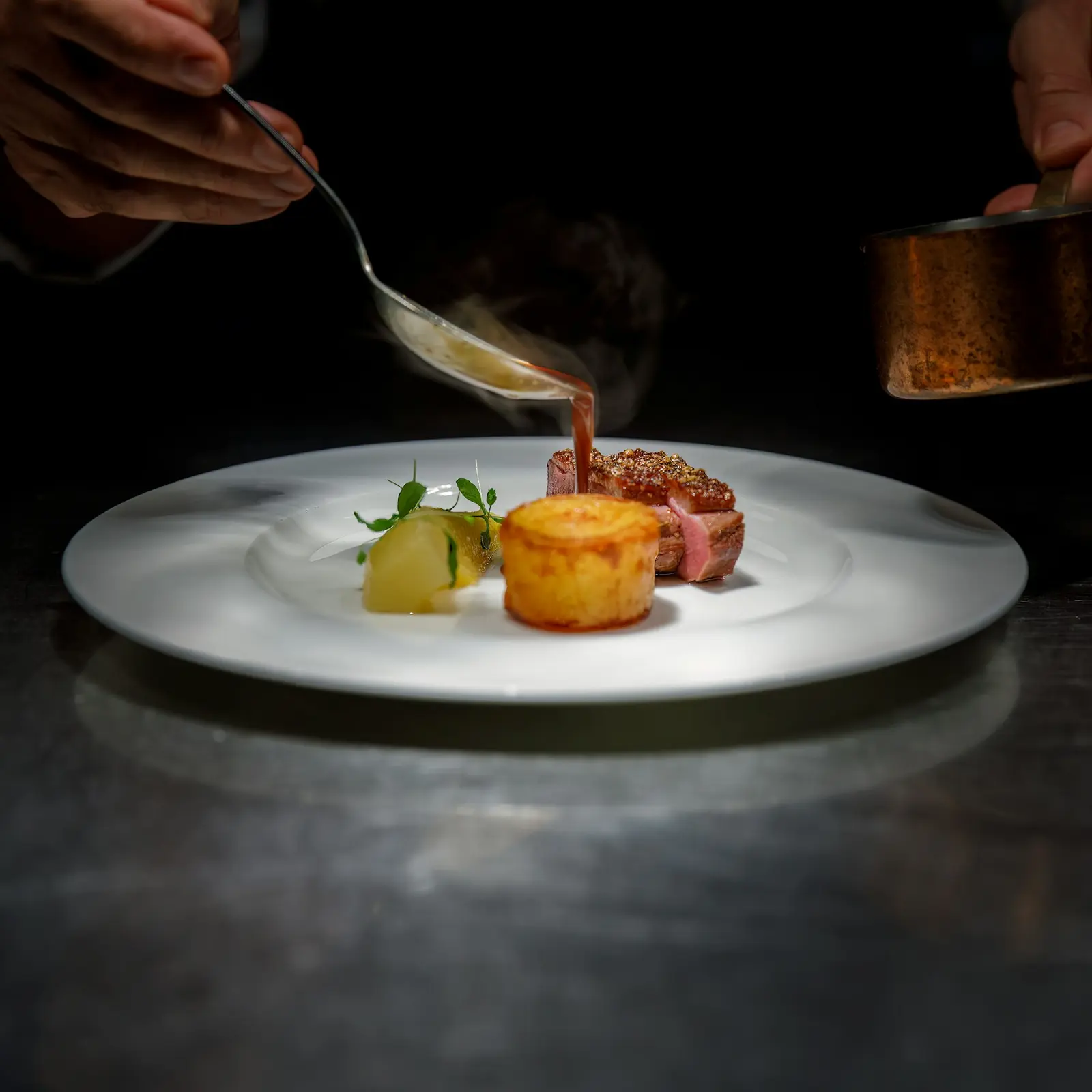A chef spoons steaming jus over duck breast and a golden potato pavé on a white plate