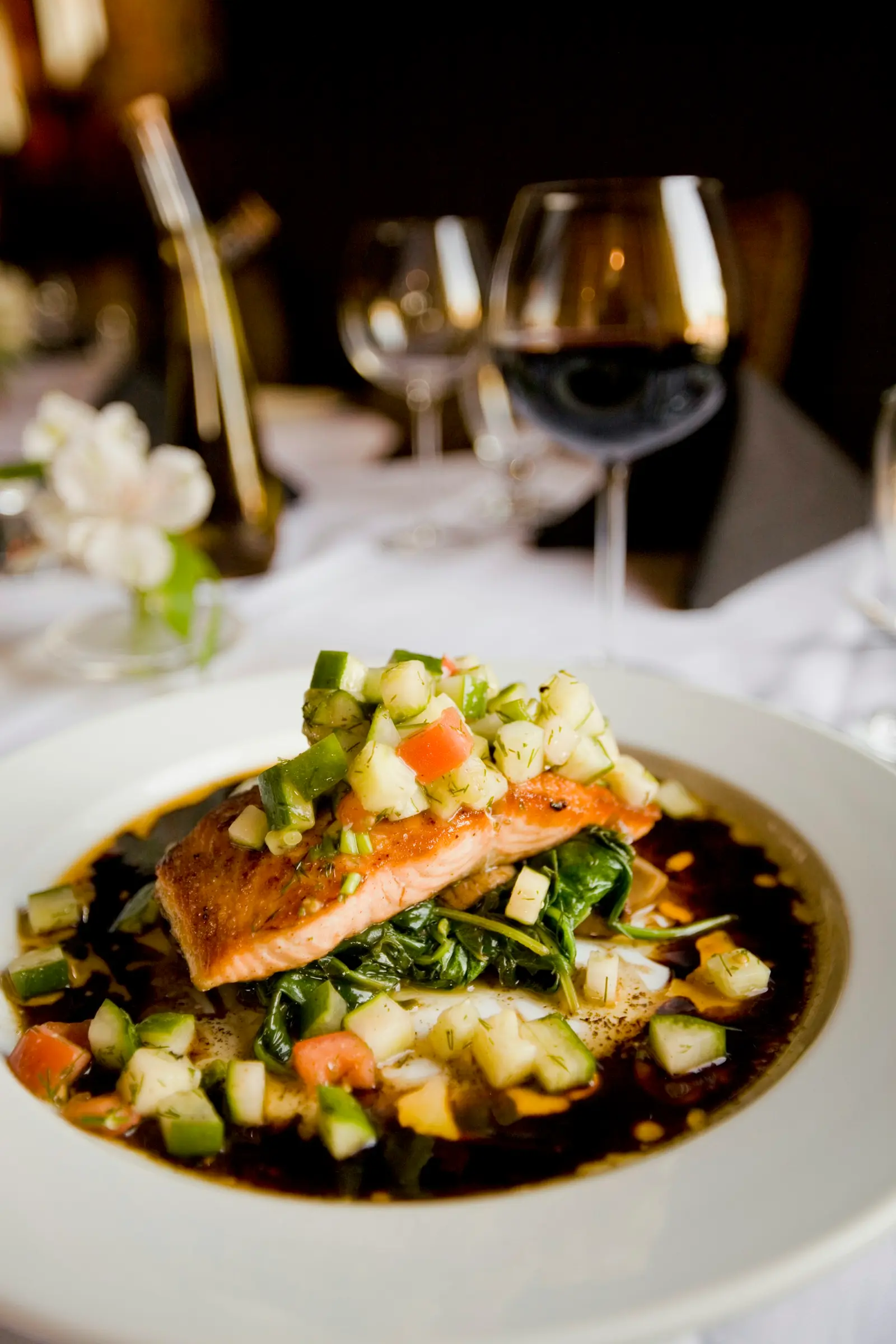 Seared salmon over wilted spinach on a white plate in a dark dining room, a glass of red wine behind