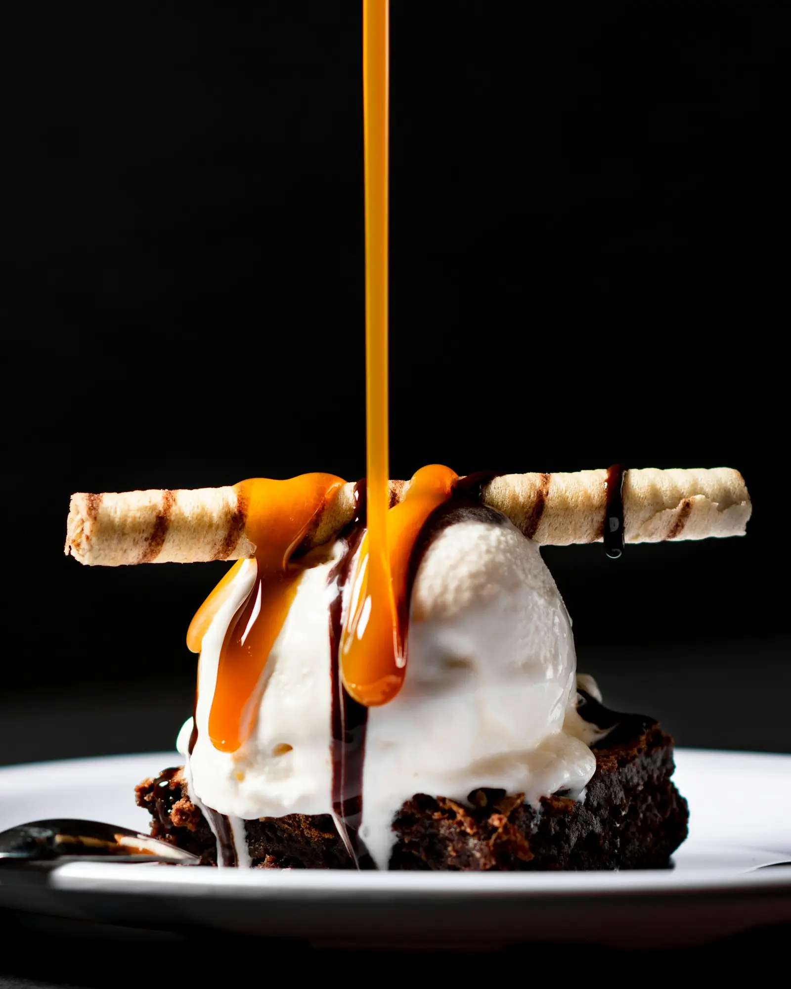Caramel poured in a thin ribbon over ice cream and a dark chocolate brownie against a black background