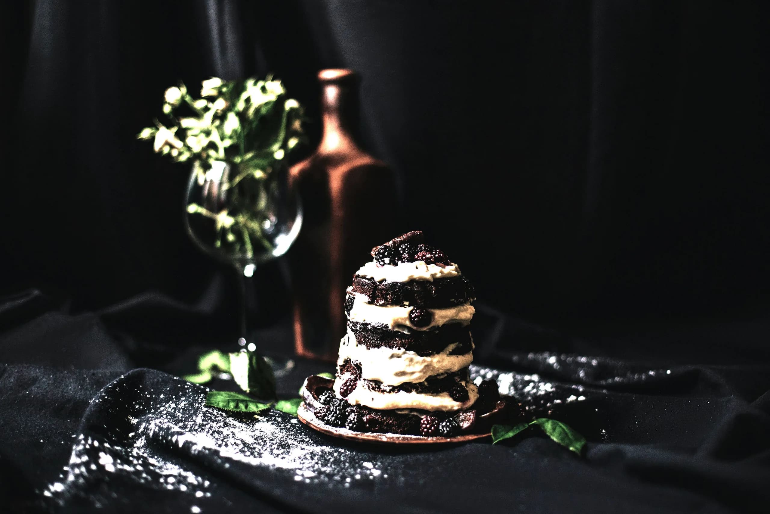 A stacked dark chocolate and cream dessert with blackberries on a dark cloth dusted with sugar