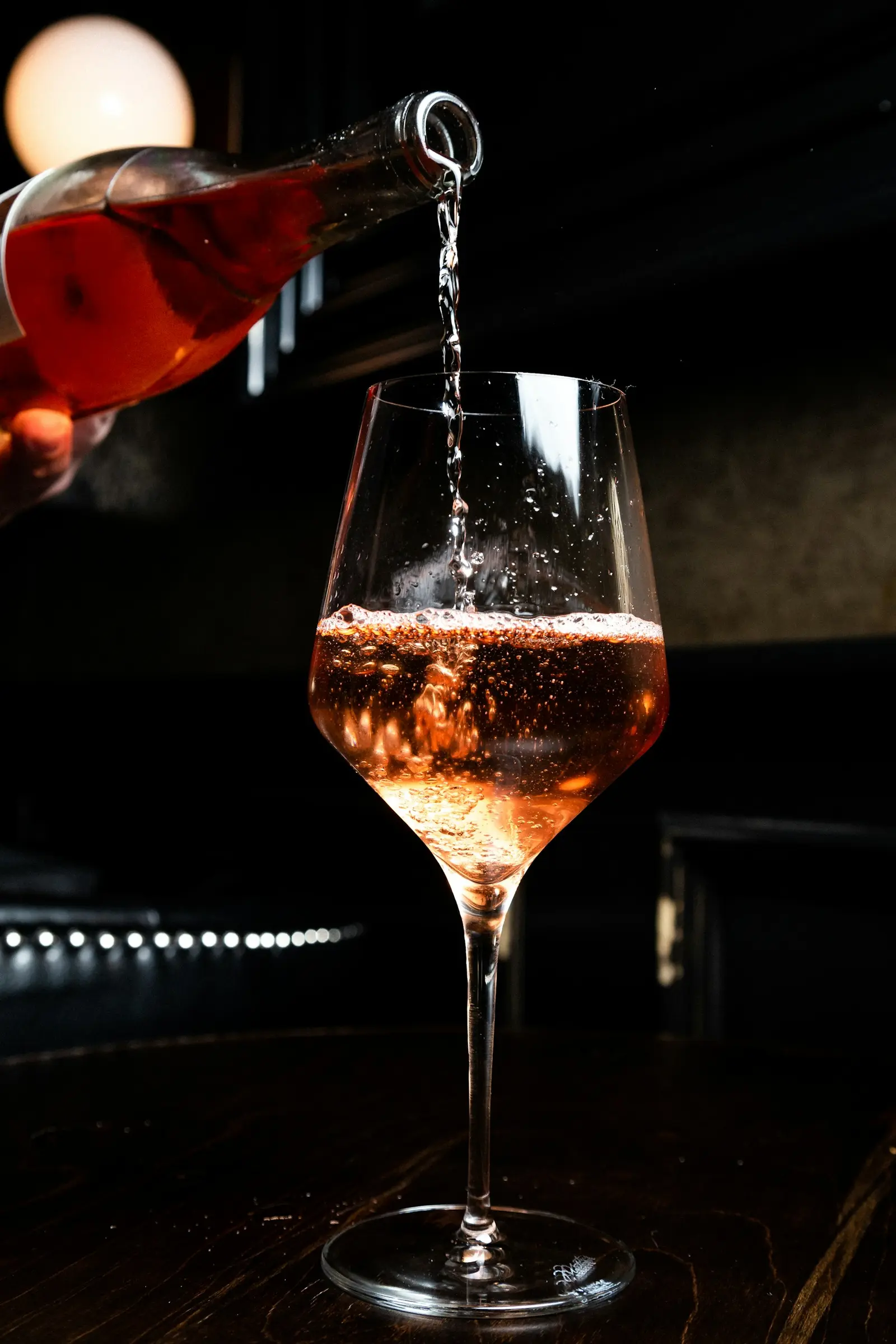 Rosé wine being poured into a stemmed glass against a dark bar