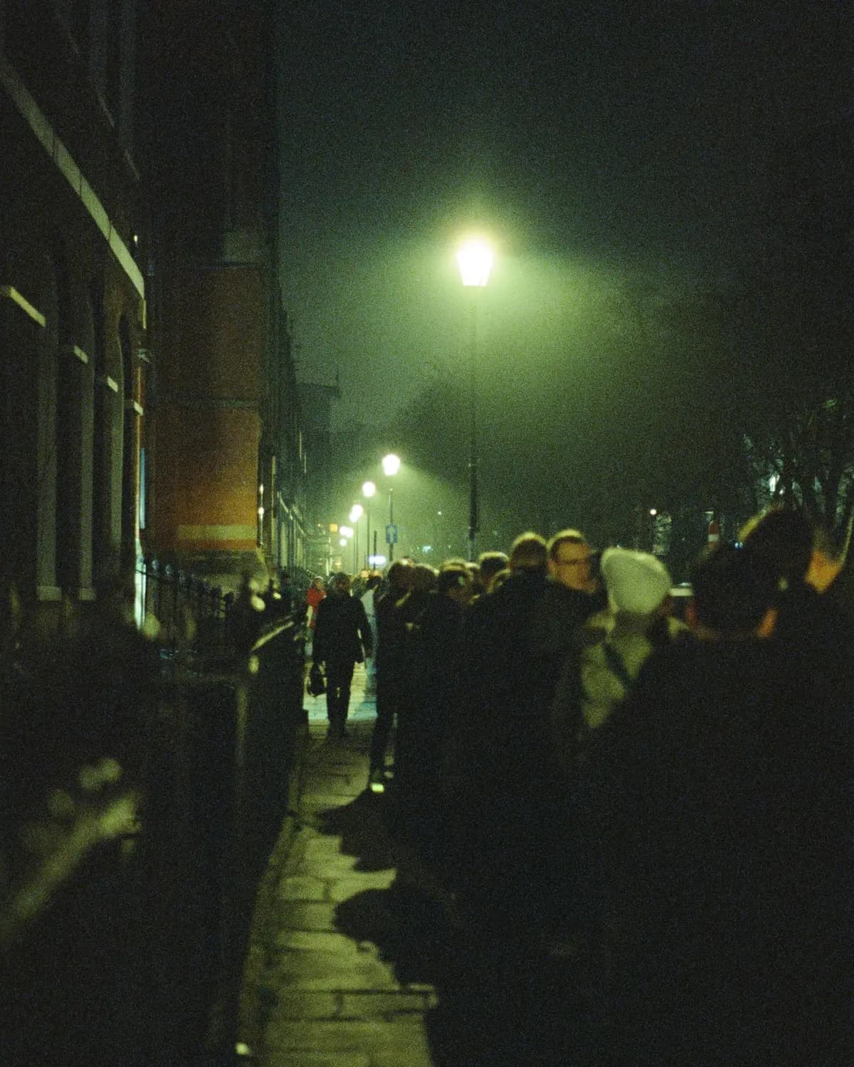 A queue of people on a wet pavement at night, under a sodium streetlight.
