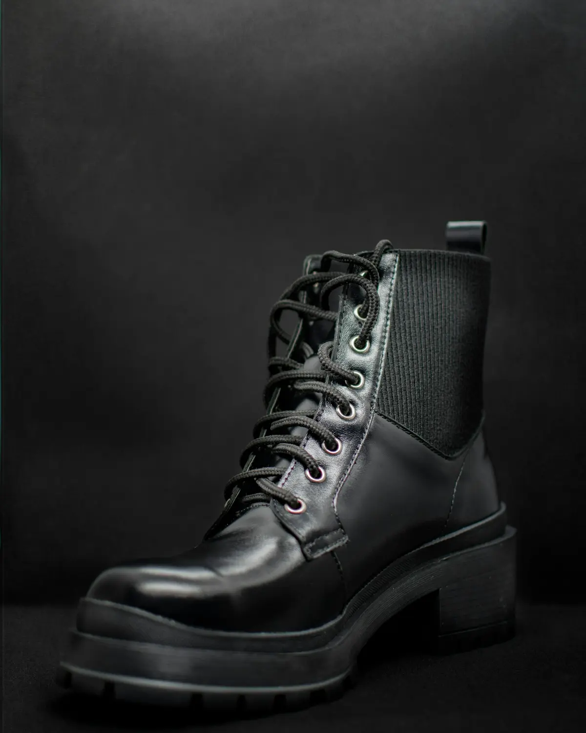 A black lace-up leather boot with a lug sole and an elastic gusset, lit against a dark studio backdrop.