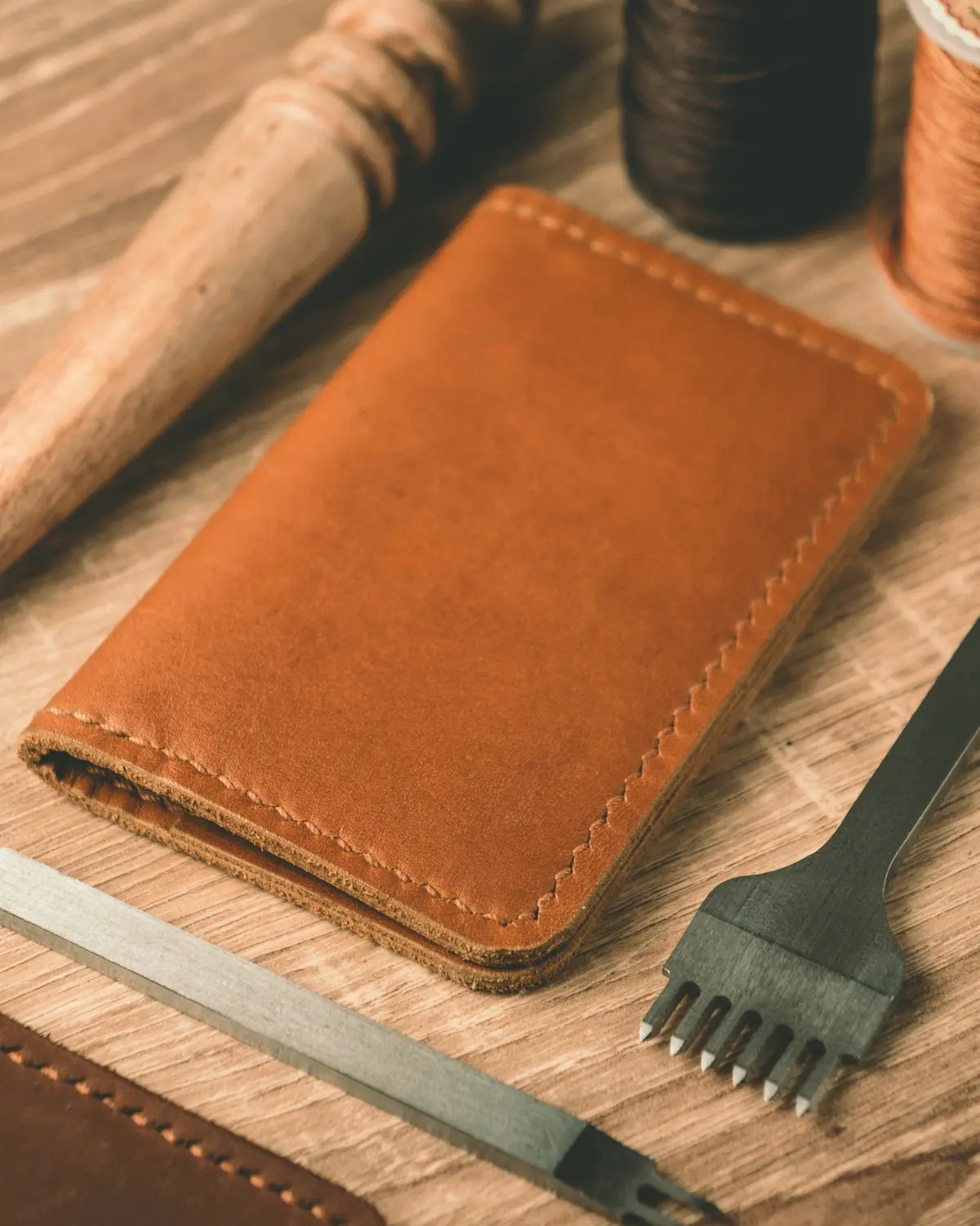 A hand-stitched tan leather card wallet on a workbench with waxed thread spools, a pricking iron and an edge tool.