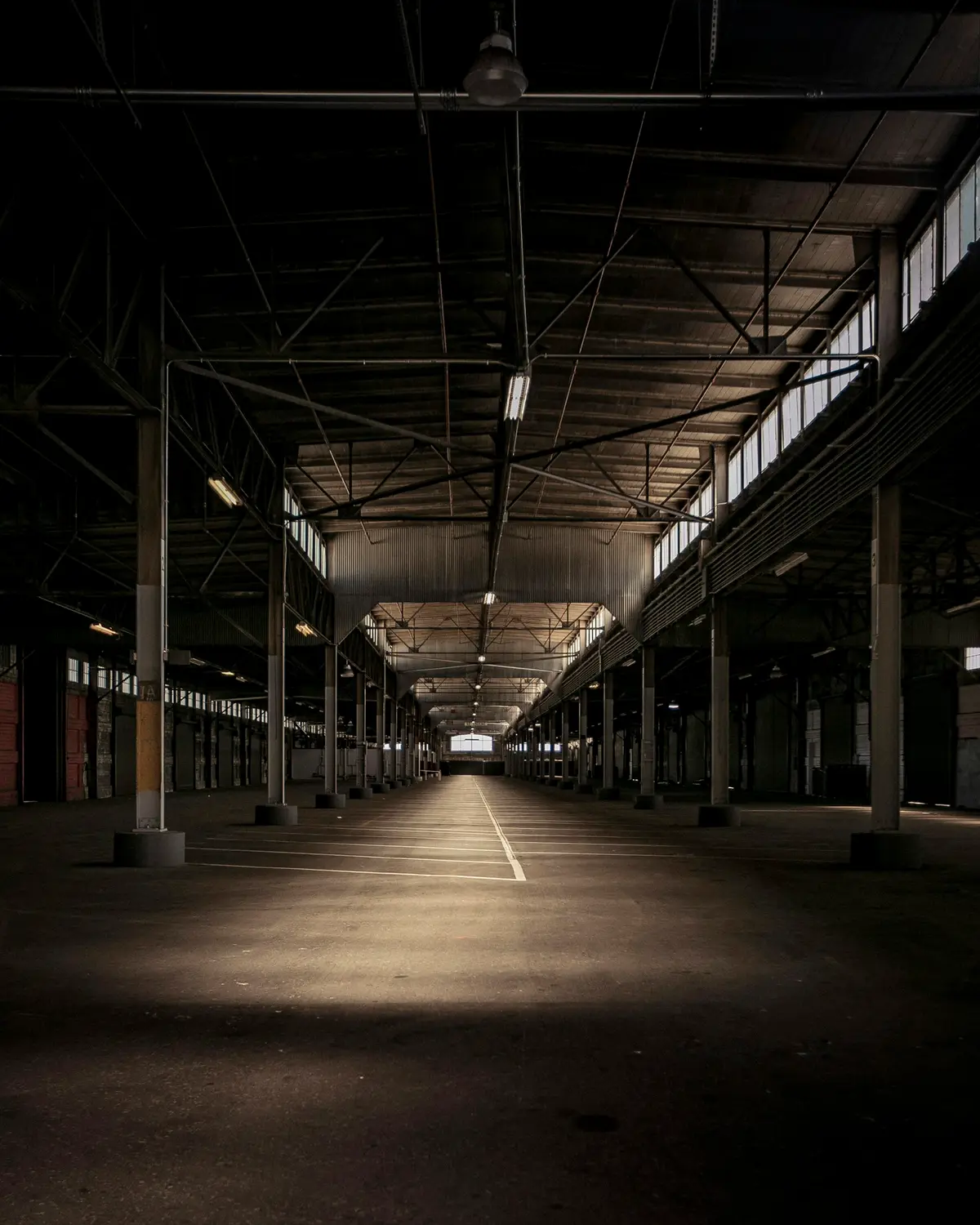 An empty industrial hall with steel columns and one lit aisle running to the far wall.