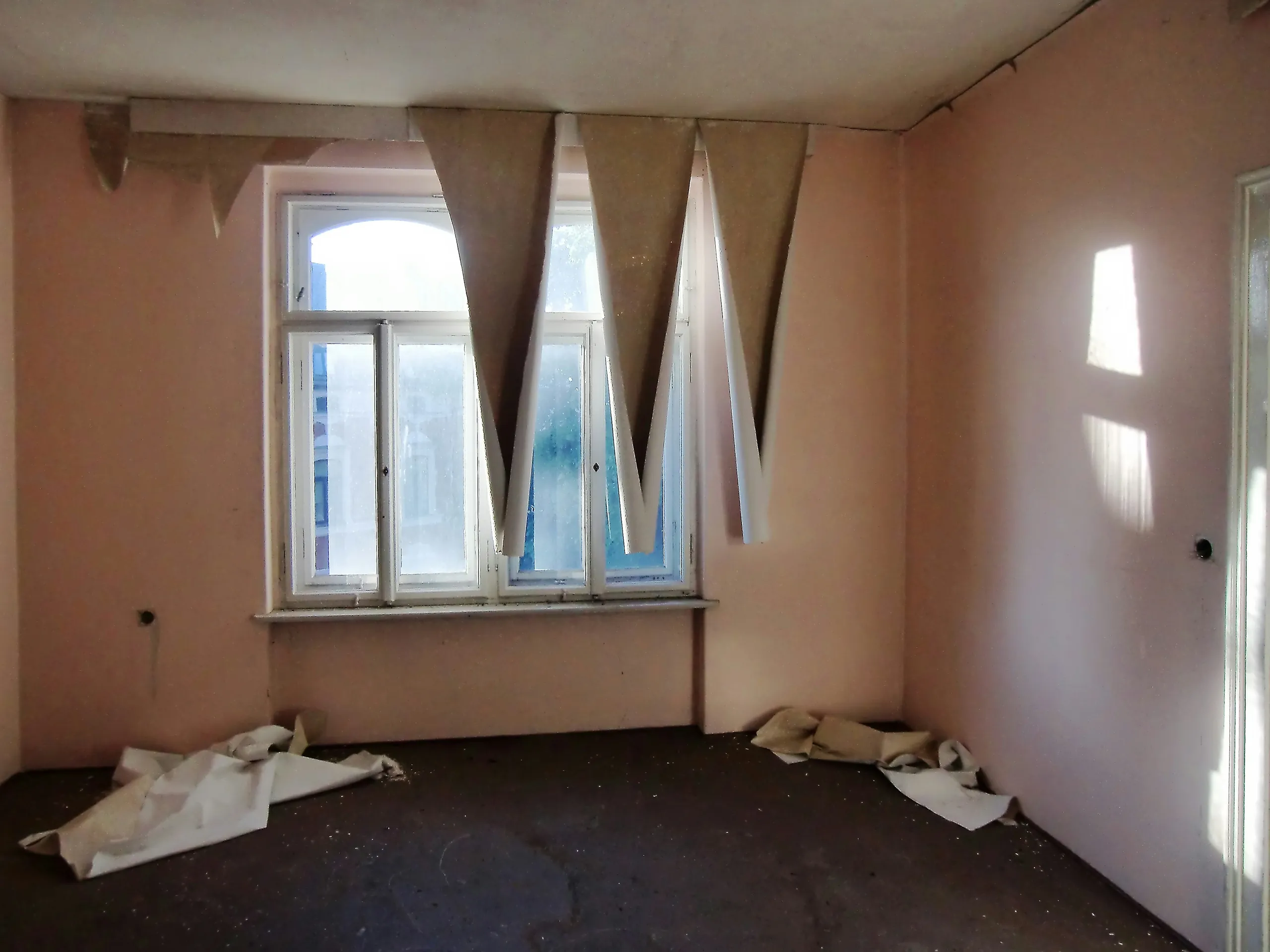 A small empty room with pink walls and torn lining paper hanging in strips over a bare window