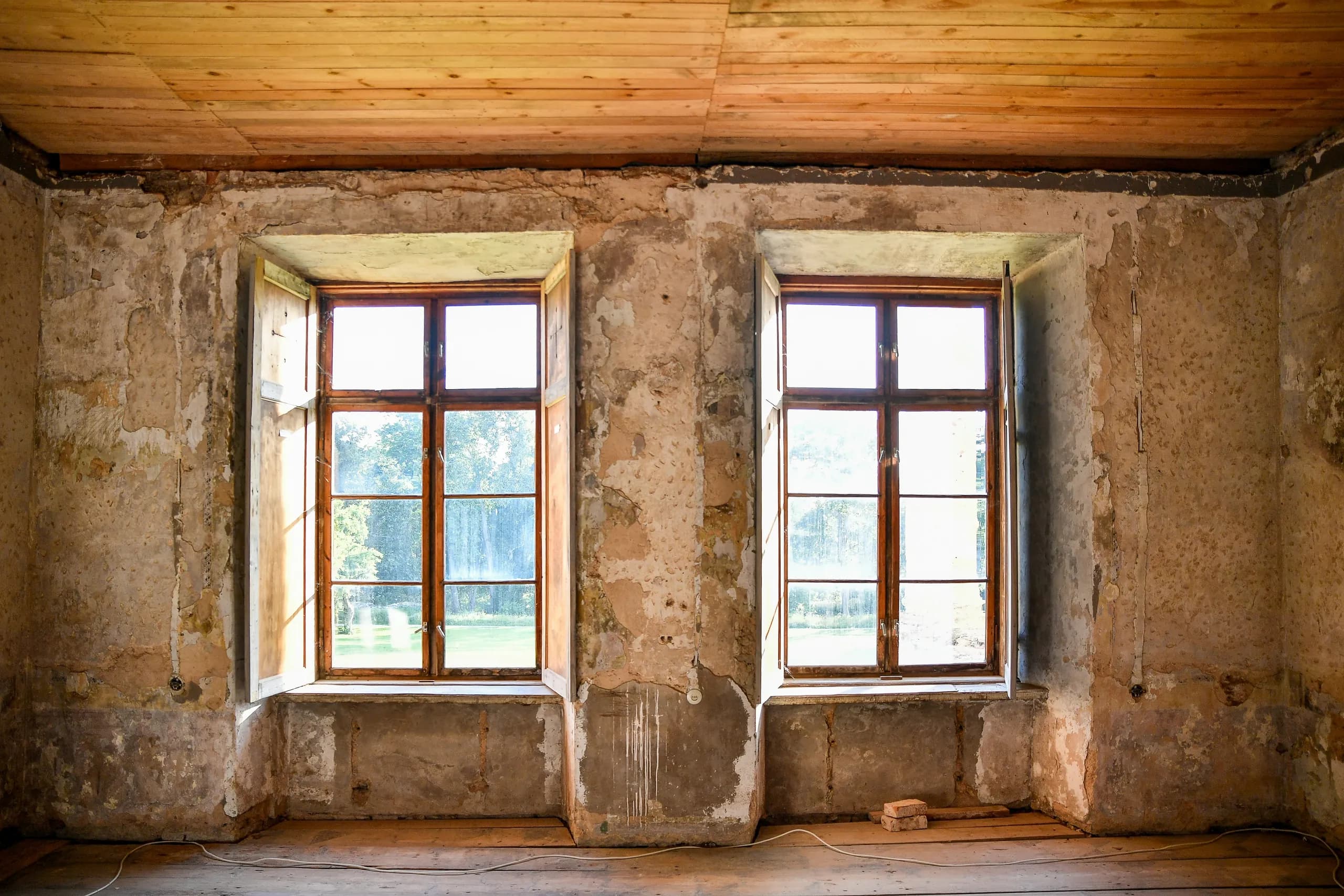 An empty period room with two tall casement windows, bare blown plaster walls and a boarded pine ceiling
