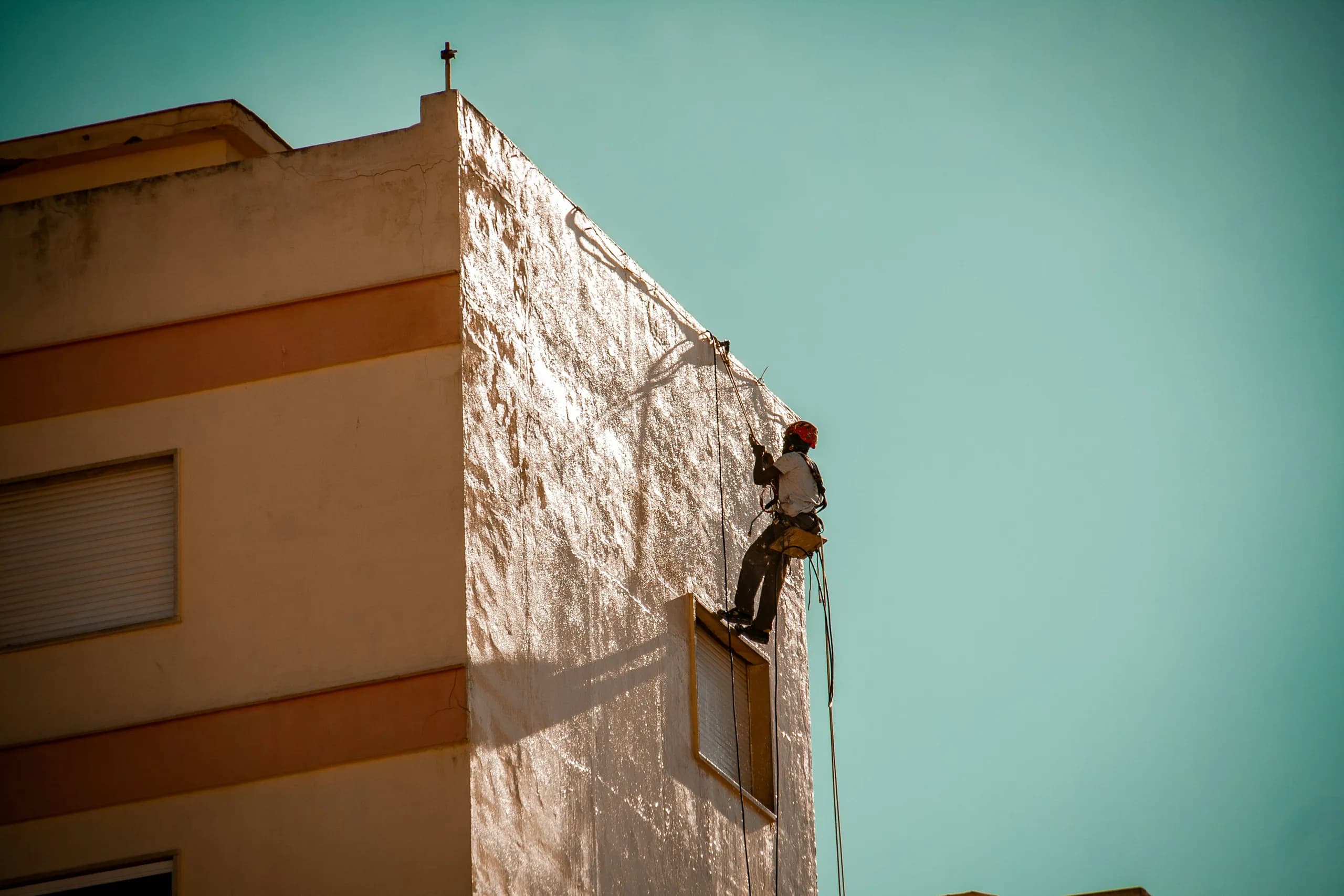 A painter on rope access working the render at the top corner of a building against a clear sky