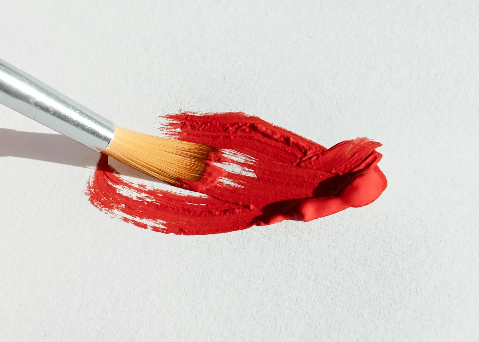 Loaded artist's brush dragging a thick stroke of vivid red-orange paint across white paper