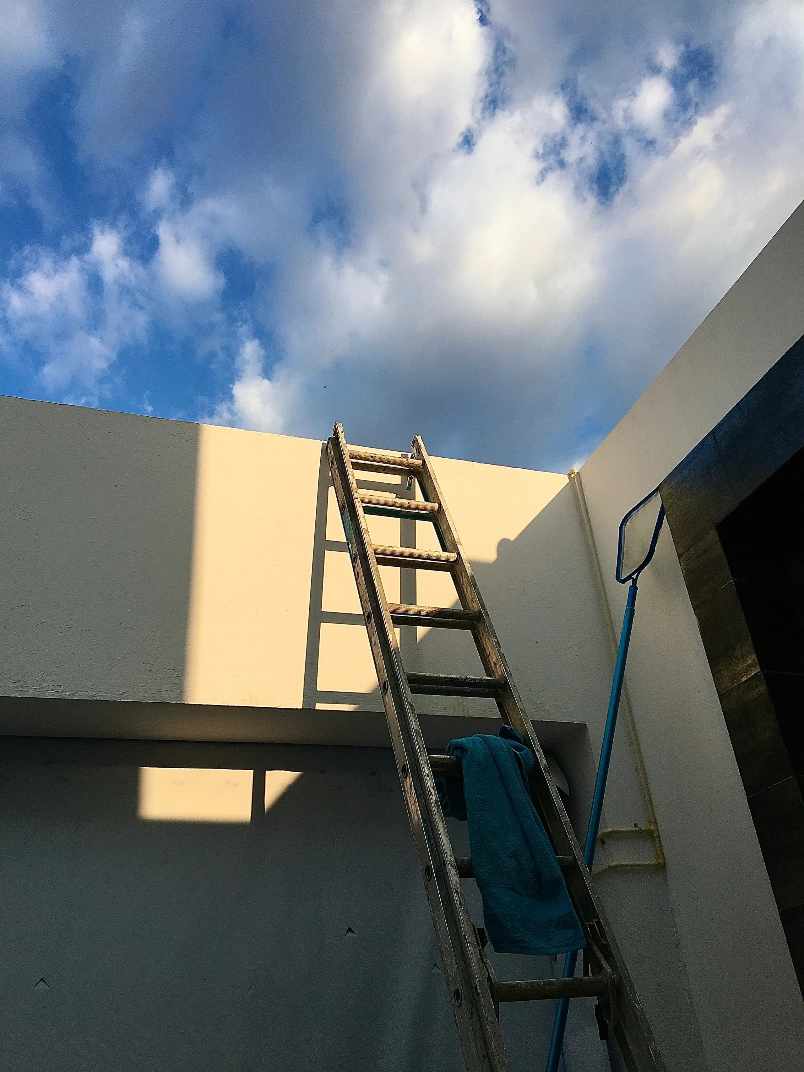 A wooden ladder leaning against a cream rendered parapet wall under a bright sky