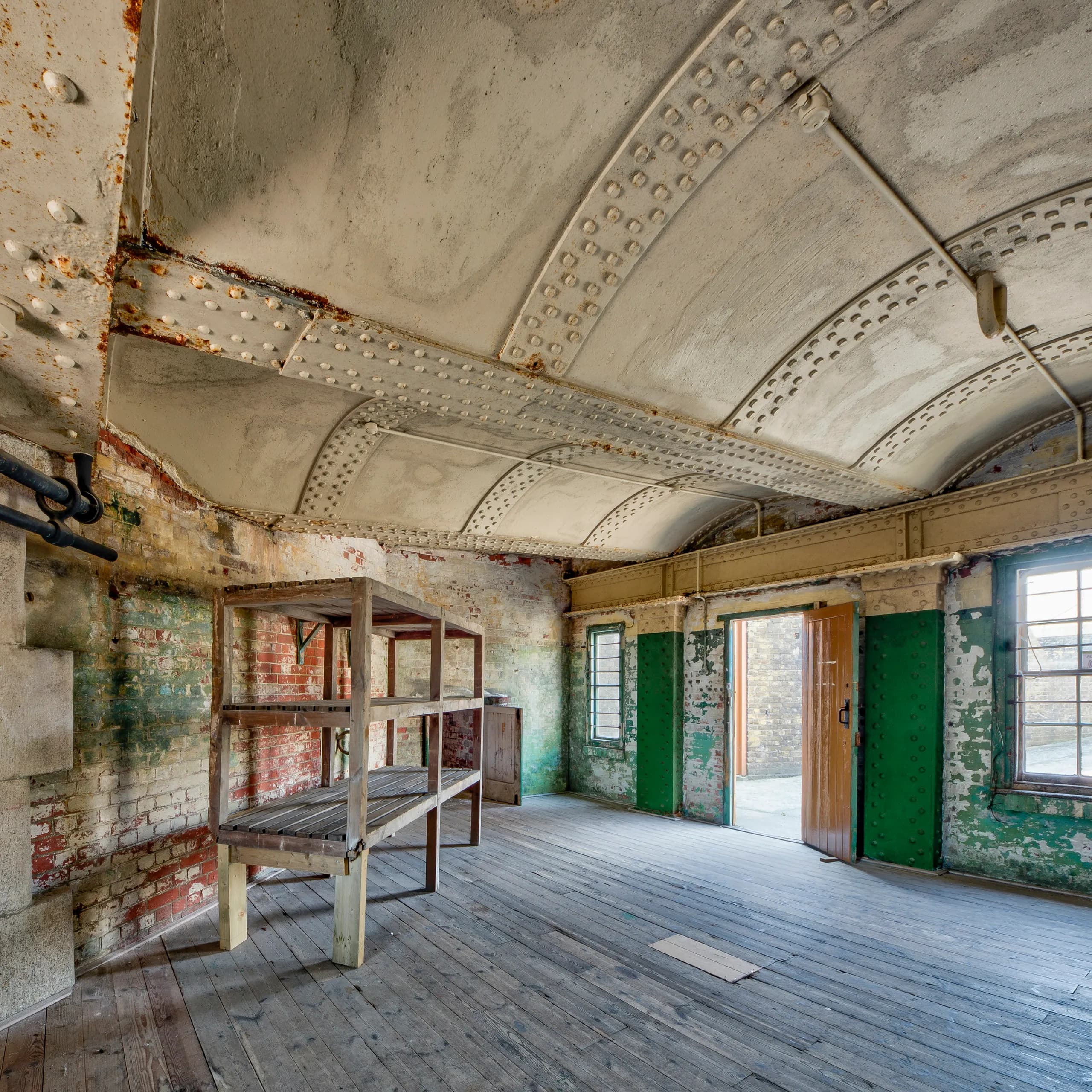 A stripped industrial room with a riveted vaulted ceiling, distressed brick walls and green iron doors