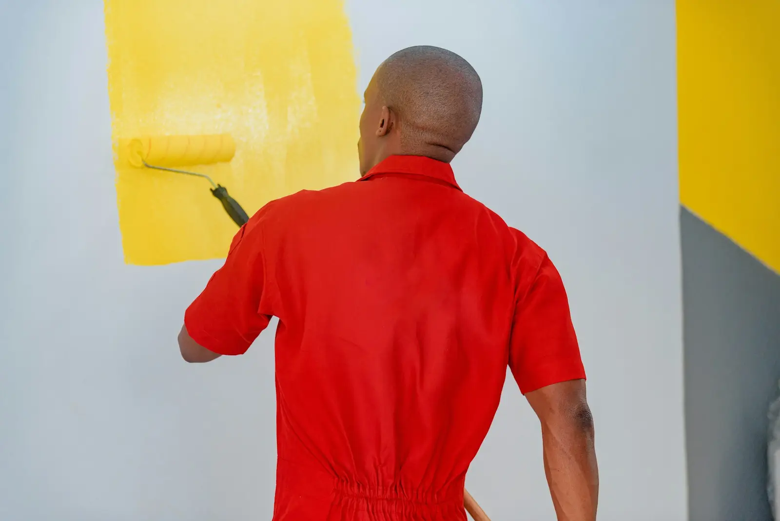 Painter in red coveralls rolling bright yellow paint onto a pale wall