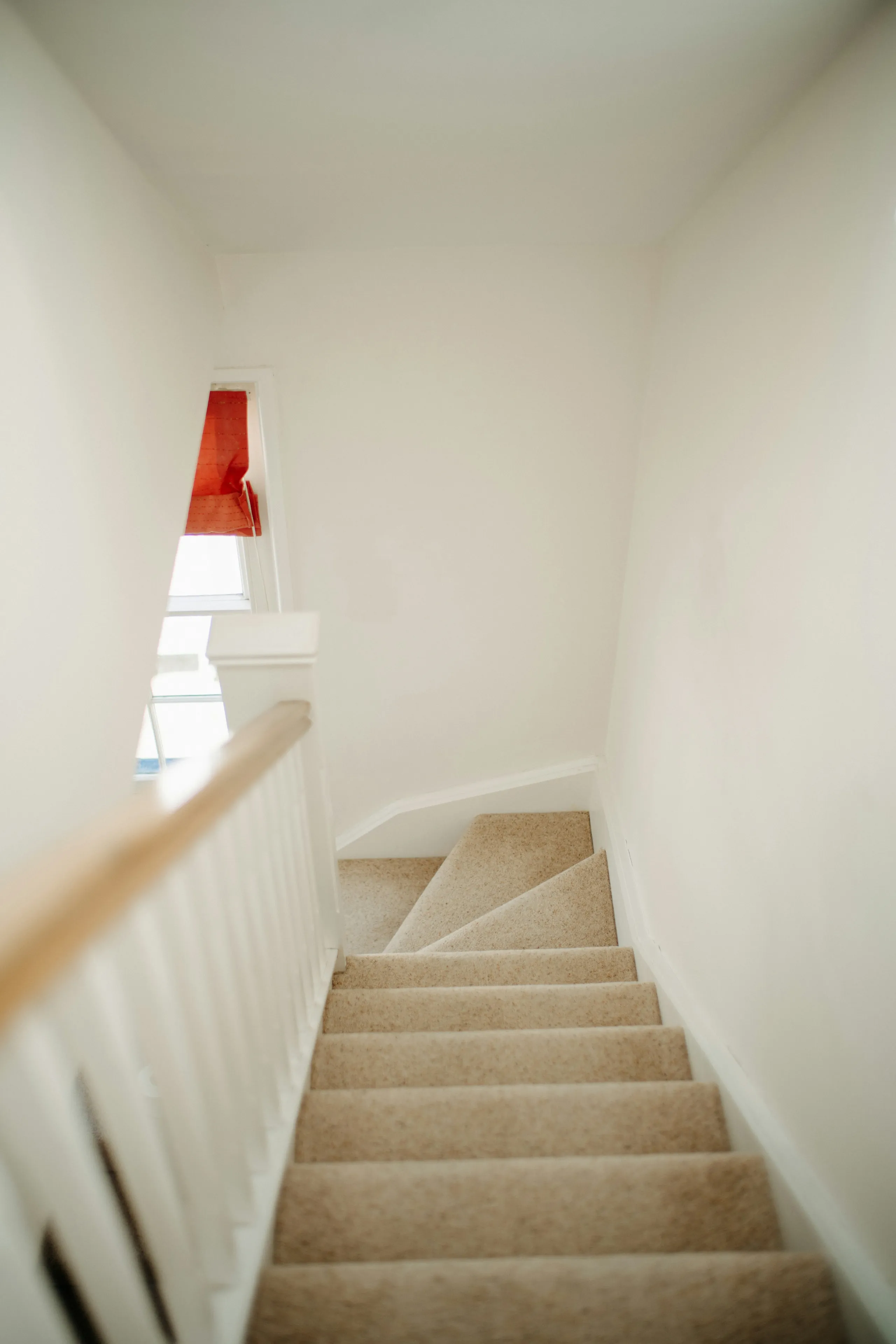 A narrow stair hall painted flat off-white, looking down the treads past a window with a red blind