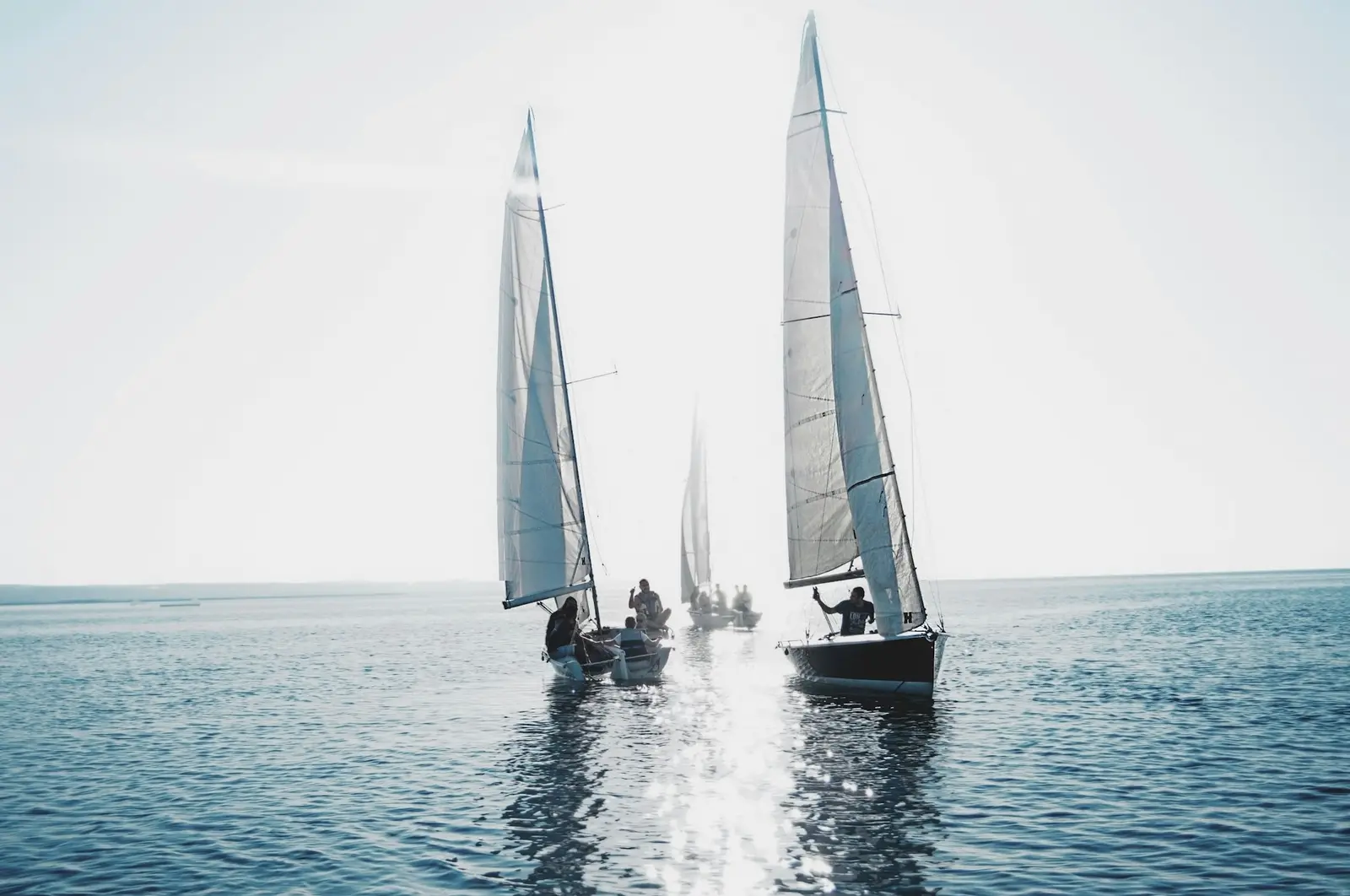 Two matched one-design keelboats sailing side by side under white sails, a third astern, backlit on flat water
