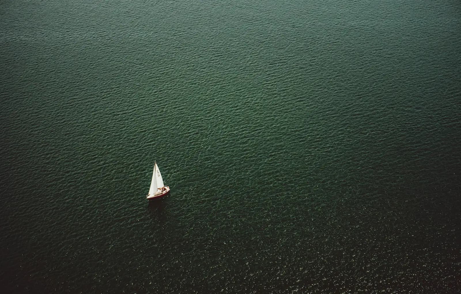 Aerial view of a lone white sloop crossing deep green open water under main and jib
