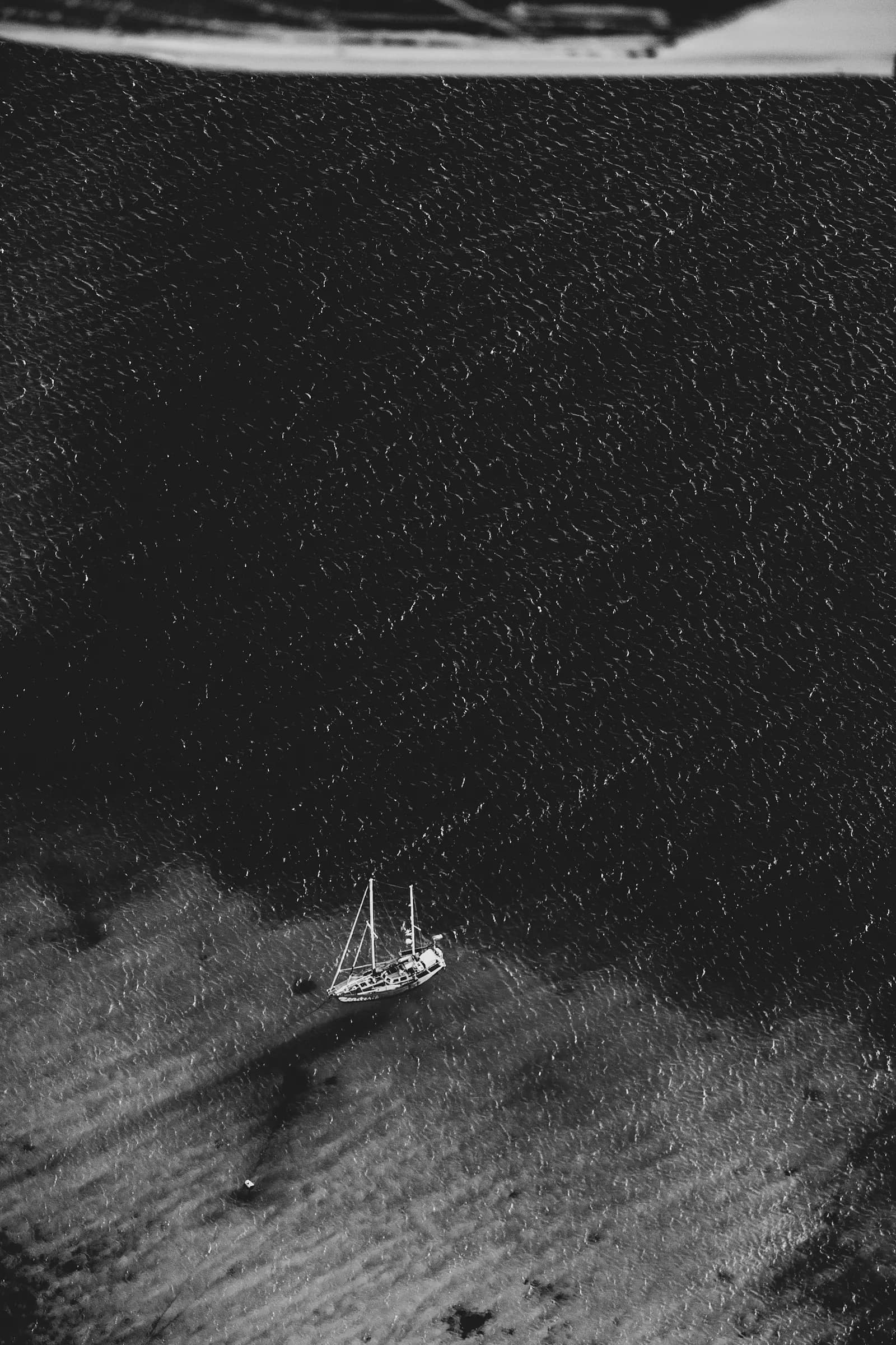 Black-and-white aerial of a two-masted yacht anchored over a pale seabed