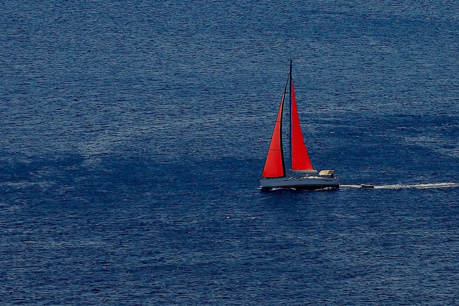 Sloop under red main and genoa crossing deep blue open water, a tender towed astern
