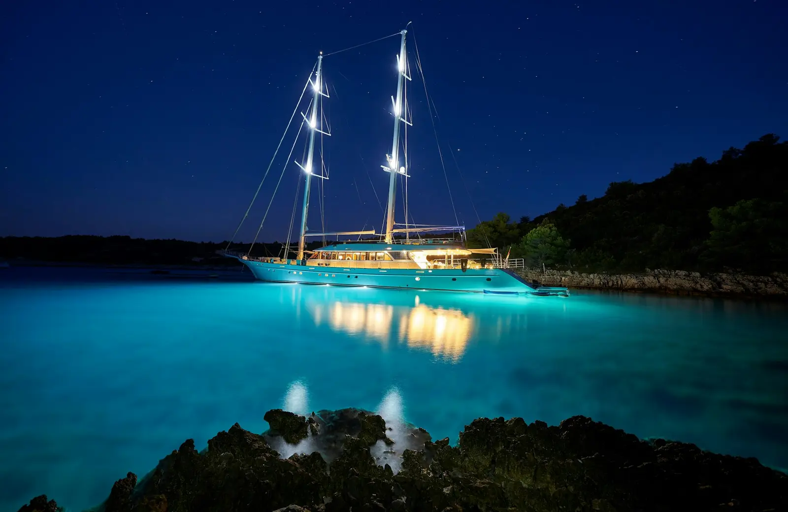 Two-masted sailing yacht lit up at anchor in a dark bay, its lights glowing on turquoise water